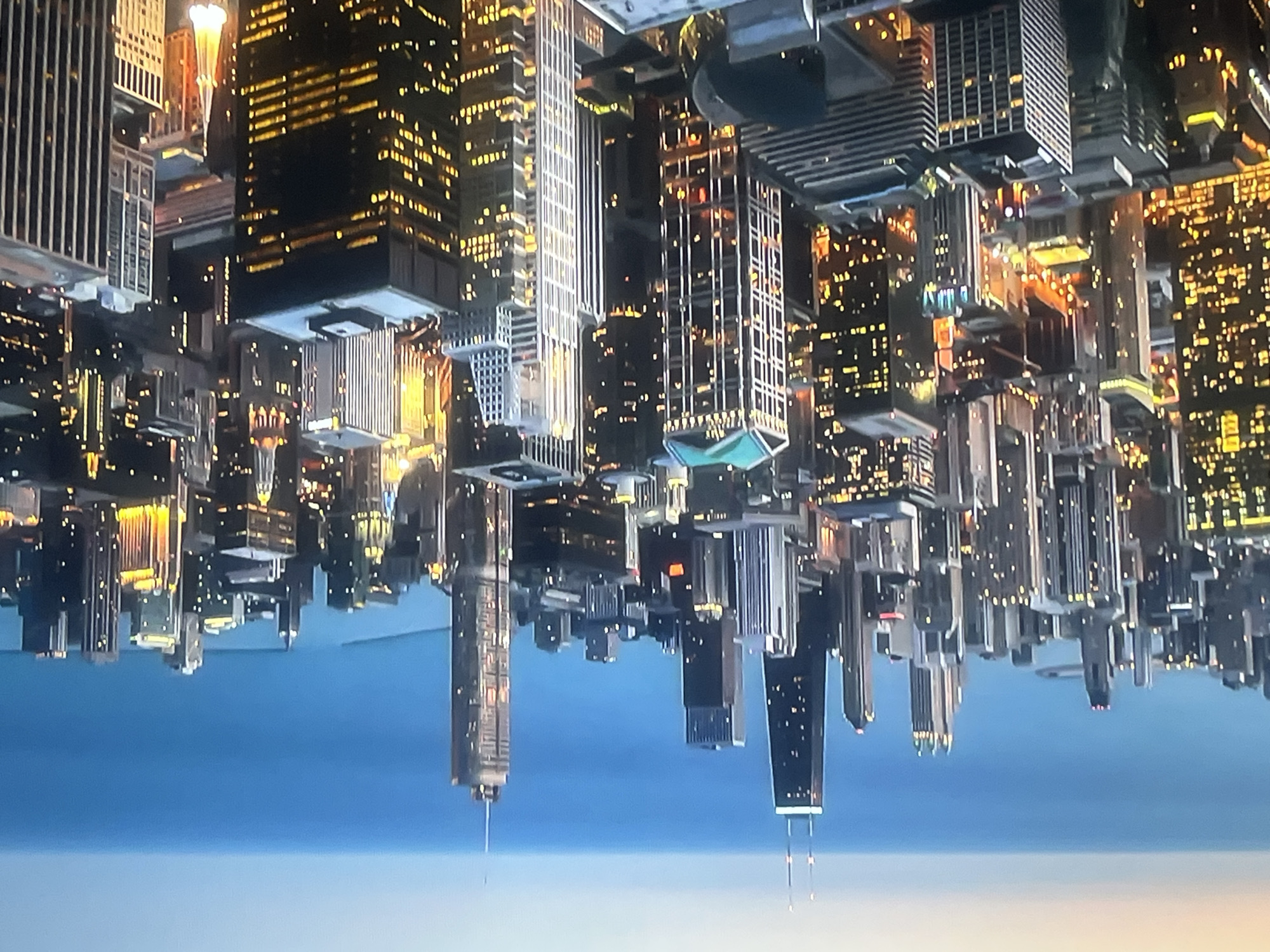 An aerial, high-angle, long shot shows a dense urban cityscape at dusk, with a vast body of water in the background. The sky transitions from a pale orange and pink near the horizon to a deep blue overhead. The city skyline is dominated by numerous skyscrapers of varying heights and architectural styles. Many buildings are illuminated with yellow and white lights, indicating that it is nighttime or late evening.
In the foreground, several prominent skyscrapers are visible. One tall, dark skyscraper with two antenna-like structures on top stands out. To its right, another skyscraper with a slender, rectangular shape is illuminated by numerous lit windows. Further into the city, a building with a distinctive green-capped roof is visible, as are other structures with more modern, geometric designs. The buildings are packed closely together, creating a complex, layered effect.
The water in the background appears calm and stretches across the horizon, with a faint, lighter line suggesting a distant landmass or the curve of the Earth. The overall impression is one of a sprawling, vibrant metropolis at the edge of a large body of water. There are no people visible in the image.