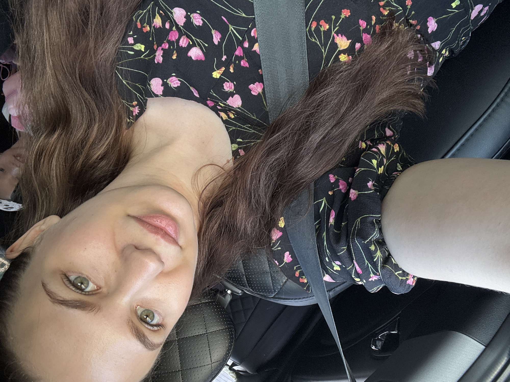 The media file is a selfie taken by a woman in a car. The woman has long, brown, wavy hair and green eyes. She is wearing a black floral dress. A gray seatbelt is visible across her chest. The background shows the interior of a car, including black leather seats. The lighting suggests it is daytime. The woman appears relaxed and is looking at the camera. No text is visible in the media file.