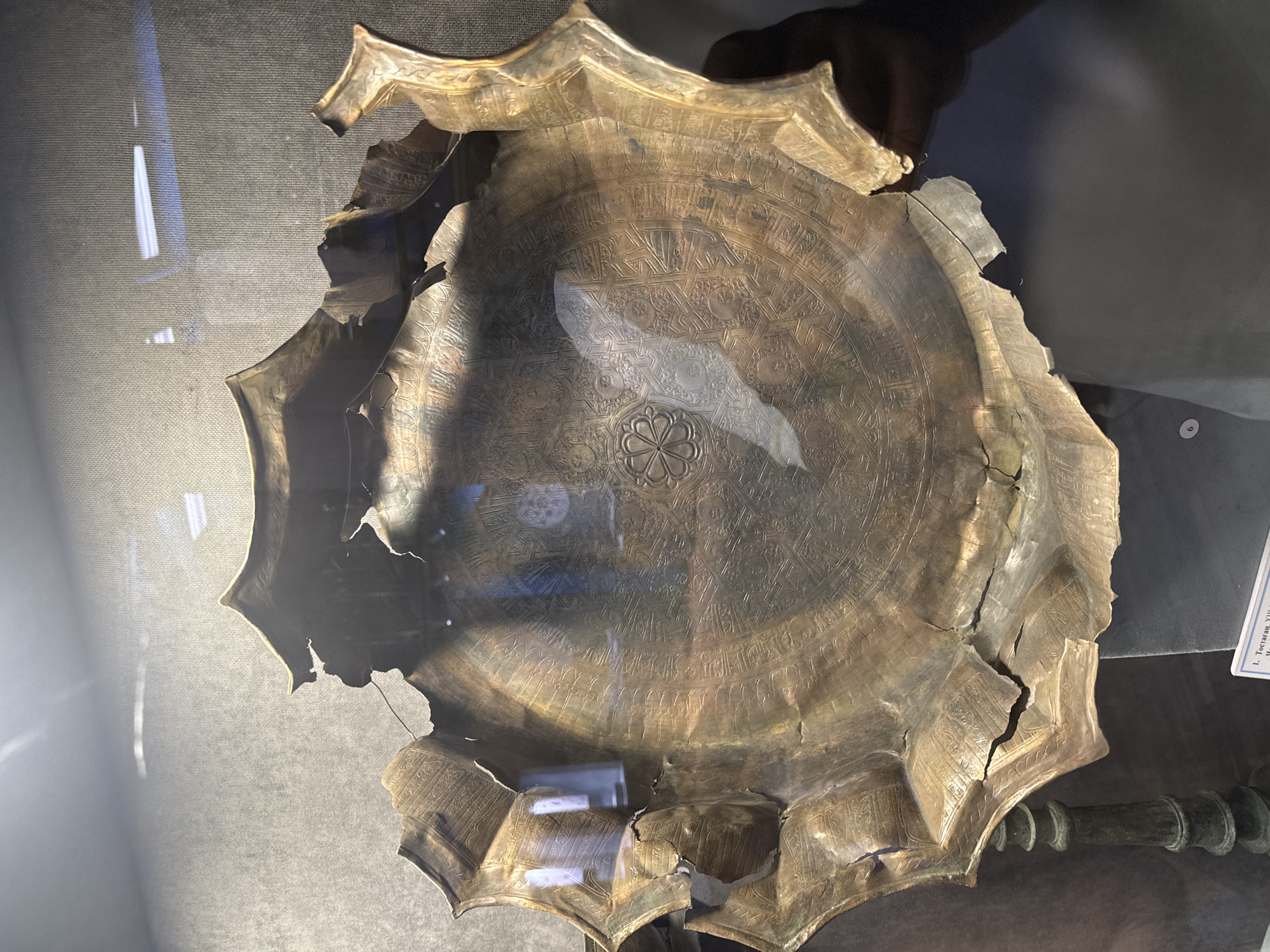 An antique, damaged, ornate metal dish is displayed in a museum in Almaty, Kazakhstan. The dish, likely made of bronze or a similar metal, has a complex geometric pattern engraved in its center, with a floral motif at the very core. The edges of the dish are broken and uneven, showing signs of age and significant damage. The dish is supported by a decorative metal stand. The background is a dark, textured wall, and there is a glass barrier in front of the dish, which reflects some ambient light and hints of figures or objects in the surrounding environment. A label with Cyrillic text is partially visible at the bottom of the image.