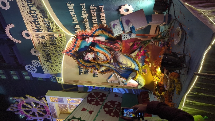 The image displays a Saraswati Puja celebration. The statue of the goddess Saraswati, adorned with garlands, is placed against a backdrop with writings in Bengali. The background includes the words "Robotics & Engi", "Mechatron Engineering", "Phule learning", "knowledge", "curiosity" and "wation". There are also decorative gears and books. The scene is lit by spotlights, and a person is taking a photo with their mobile. The entire setting seems to be indoors with detailed artistic work.