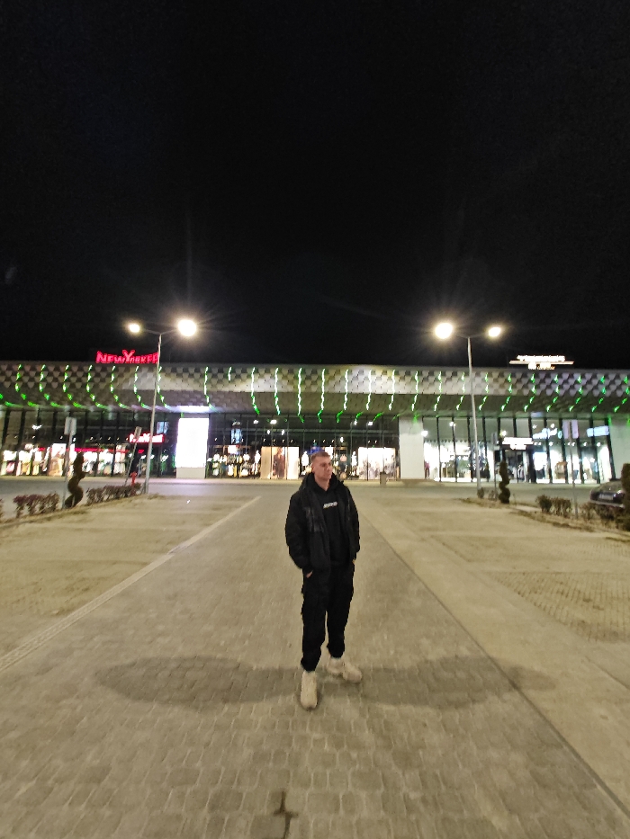 The image captures a nighttime scene at Grand Mall Shumen, Shumen, Bulgaria. A single male subject stands centrally in the foreground on a paved pedestrian walkway, facing slightly left. He is dressed in a black hooded sweatshirt with a white logo, a black open puffer jacket, black cargo pants, and light-colored chunky sneakers. His hands are in his jacket pockets.
Behind the individual, the brightly lit facade of the Grand Mall Shumen is visible, characterized by large glass windows displaying various illuminated storefronts. The upper section of the building features a distinctive black and white checkered pattern accented by horizontal lines of vibrant green LED lights. A prominent red "New Yorker" sign is affixed to the upper-left portion of the facade. The area is illuminated by multiple streetlights featuring dual lamps, with one positioned directly behind the subject. The sky above is dark and clear.