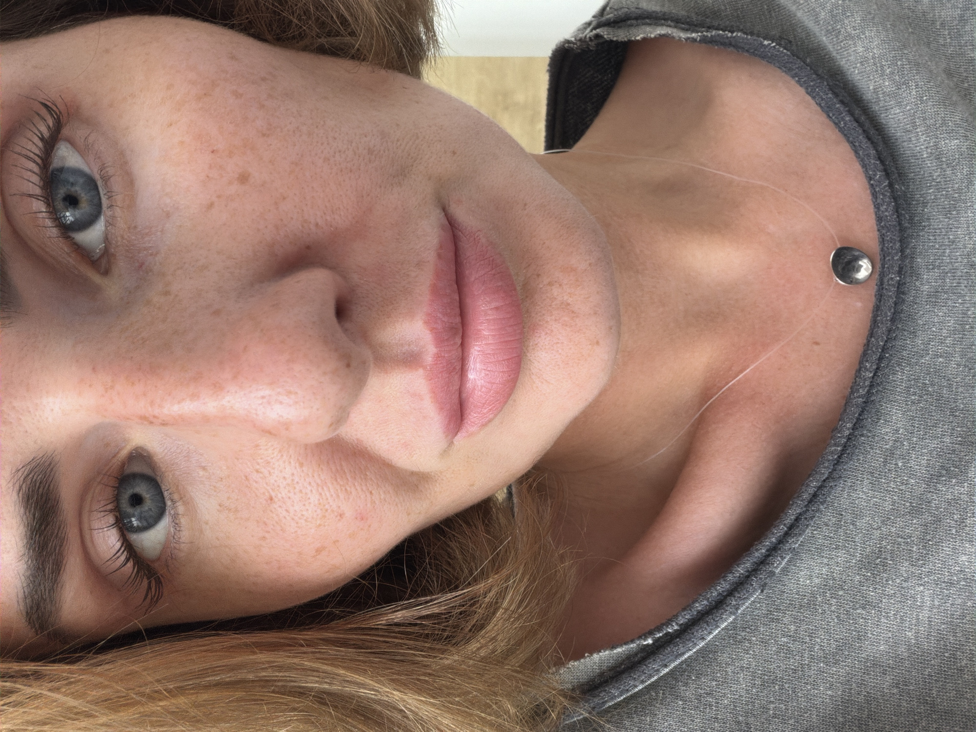 A close-up image shows a woman with fair skin, freckles, and blue eyes. Her hair is light brown. She is wearing a gray top and a necklace with a silver pendant on a transparent cord. The image is oriented sideways. The background is out of focus. There is no indication in the image or description that the image was taken in Bac Nha Trang Ward, Vietnam.