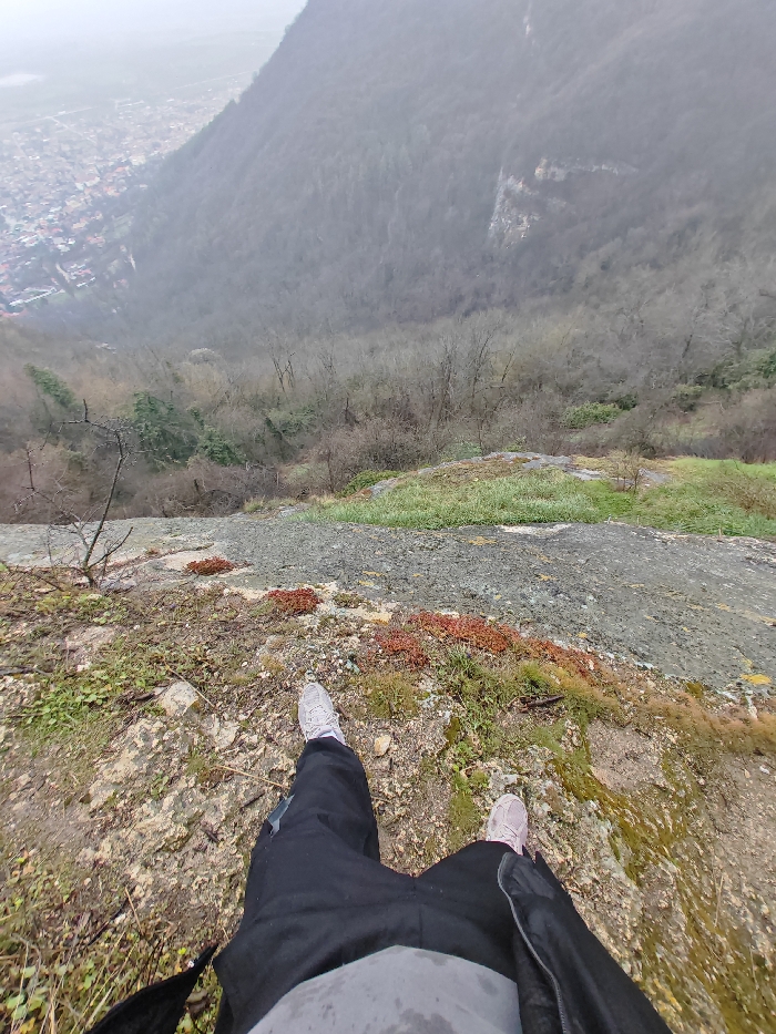 The image depicts a downward-angle view from a precipitous rocky outcrop on the Shumen Plateau, overlooking the city of Shumen, Bulgaria, and the surrounding forested valley. A person, visible from the torso down, stands on the edge of the rocky ledge, wearing black trousers, a light grey shirt, and light-colored athletic shoes. The immediate foreground consists of uneven, light-colored rock and soil, interspersed with patches of sparse green grass and reddish-brown low-lying vegetation. Beyond the ledge, a steep, densely forested mountainside descends into a valley. The trees are predominantly deciduous and bare, suggesting a non-summer season. In the distance to the left, the city of Shumen is visible, characterized by numerous buildings and urban development. The sky is overcast and hazy, creating a muted light condition.
