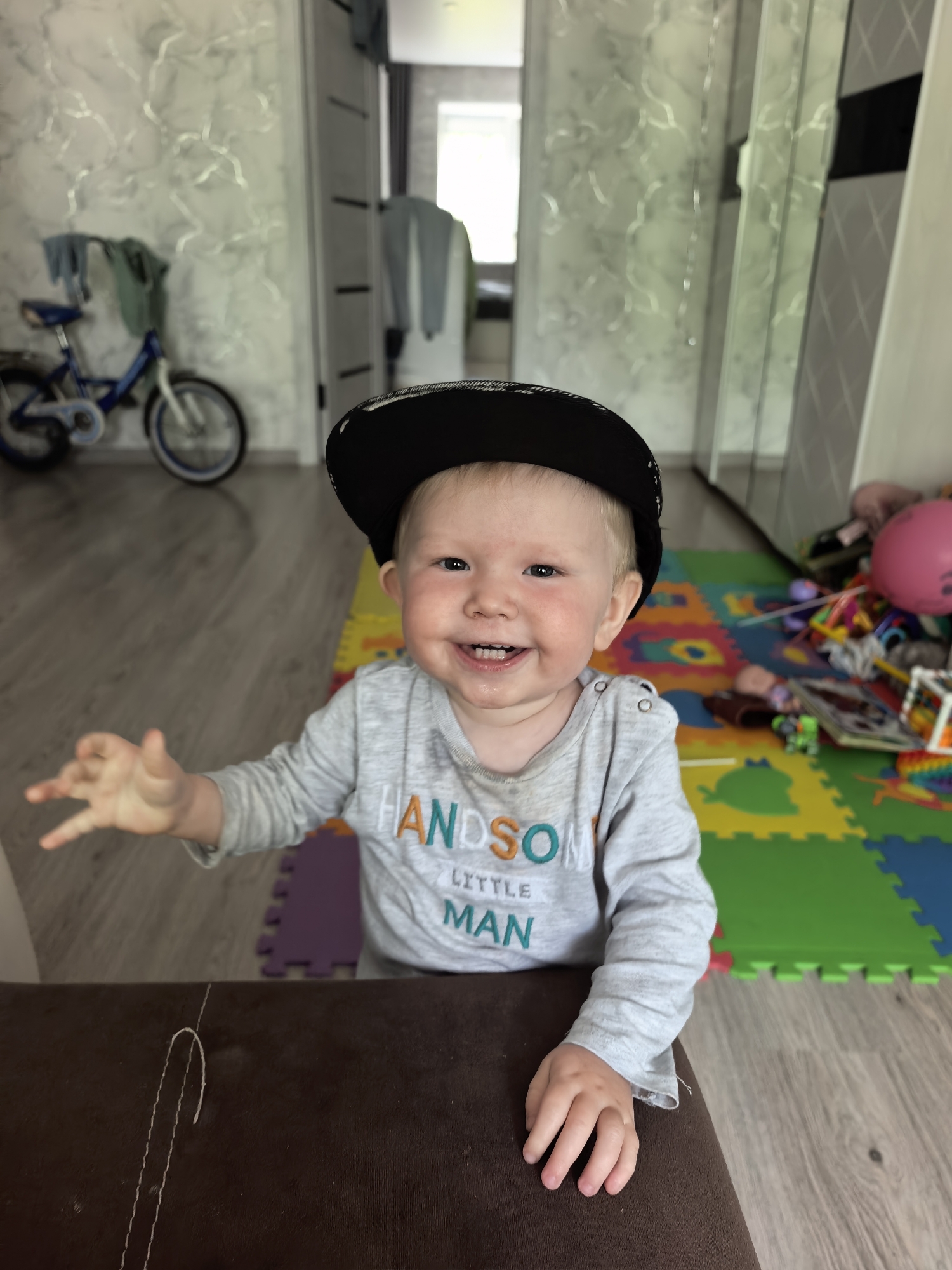 This image depicts a young child, likely a toddler, positioned in an indoor residential setting in the city of Sterlitamak, Russia. The child, with fair skin and light hair partially concealed by a backward-facing black baseball cap, is seen from the chest up, looking directly at the viewer. A wide, joyful smile reveals several visible teeth, and the child's cheeks exhibit a flush of pink. The child is wearing a light grey long-sleeved shirt with the text "HANDSOME LITTLE MAN" printed in colorful, varied fonts across the chest. The left hand is raised with fingers splayed, while the right hand rests on a dark brown, upholstered surface that appears to be part of a sofa or armchair, positioned in the lower foreground.
The background shows a brightly lit room. To the child's left (viewer's right), colorful interlocking foam playmats in shades of green, yellow, orange, and purple cover a portion of the floor, where a cluttered assortment of toys, including a deflated pink balloon, dolls, and various plastic items, is visible. To the child's right (viewer's left), the floor transitions to light-colored laminate or wood. In the mid-to-far background, a small blue child's bicycle with white wheels and fenders stands against a wall adorned with textured, light-colored wallpaper featuring subtle, wavy grey patterns. Clothes are draped over the bicycle's handlebars. Further back, light-colored doors or wardrobe panels, some with reflective surfaces, are visible, and a window allows natural light into the space.
