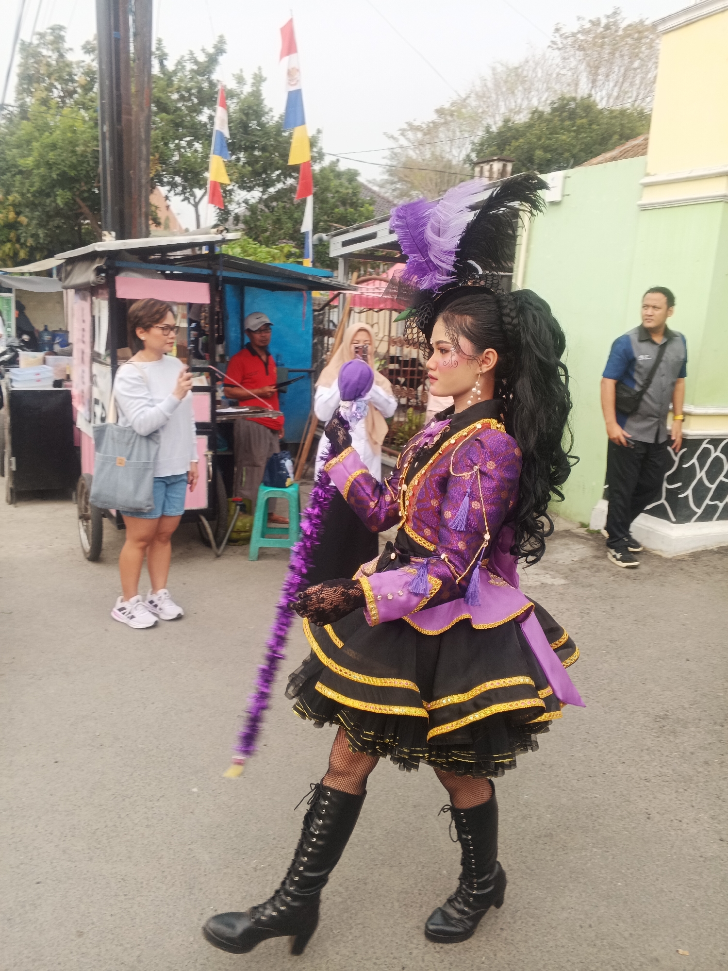 In Sokanegara, Indonesia, a young woman in elaborate purple and black attire, complete with feathers in her hair and a tall feathered staff, is the central subject of the image, captured in mid-stride on an asphalt road. She wears a short, tiered skirt decorated with gold trim and a matching jacket adorned with intricate patterns and tassels. Her legs are encased in fishnet stockings and tall, laced black boots. To her left, a woman in a white long-sleeved shirt and denim shorts, holding a phone, stands near a pink food cart. Behind her, a man in a red shirt is visible behind the cart. Further back, a man in a grey polo shirt and black pants stands near a light green building with decorative black and white patterns on its lower section. In the background, trees and a cloudy sky complete the scene, with Indonesian flags prominently displayed on poles. The overall atmosphere suggests a street event or festival.