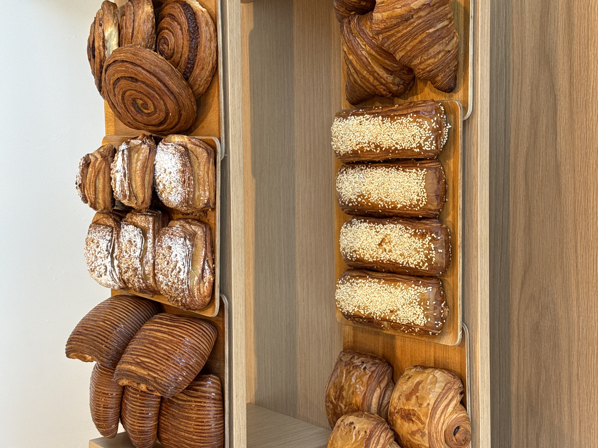 This image depicts a two-tiered display of various baked goods within a commercial establishment in Tel-Aviv, Israel. The upper shelf features three distinct varieties of pastries presented on individual wooden trays. The leftmost tray holds several large, deeply golden-brown laminated pastries with prominent ridged surfaces, resembling pain Suisse, stacked irregularly. The central tray contains multiple rectangular pastries dusted with powdered sugar and possibly nuts, consistent with pain au chocolat. The rightmost tray displays several round, spiral-shaped pastries, such as cinnamon rolls, with visible layers and some central filling, also in a deep golden-brown hue.
The lower shelf also presents three distinct groups of pastries on wooden trays. The leftmost section contains a partially visible, flaky croissant-like pastry, alongside a darker, flatter pastry and another rounded, layered item. The central tray holds four elongated, rectangular pastries, dark golden-brown and heavily coated with white sesame seeds. The rightmost tray is filled with multiple traditional croissants, characterized by their pronounced flaky layers and golden-brown exterior. All pastries are arranged on light wooden shelves, with a plain white wall visible above the top shelf. No individuals are present in the scene.