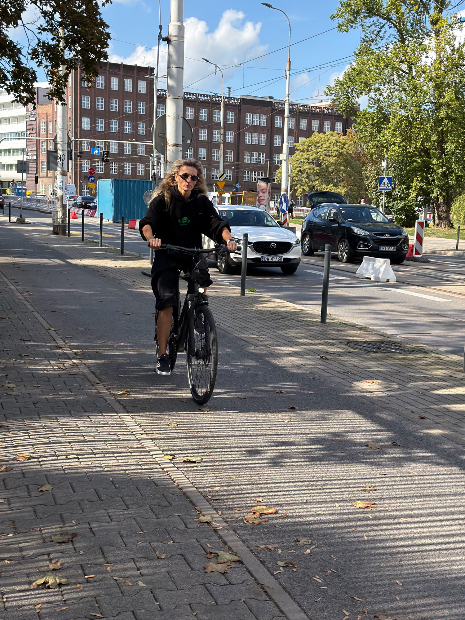 A person with long blonde hair and sunglasses, wearing a black hoodie and shorts, rides a black electric bicycle on a paved sidewalk in Wrocław, Poland. The rider is in the foreground, positioned slightly to the left of center, and is looking towards the camera. The bike has a front light and knobby tires. The sidewalk is made of grey rectangular paving stones with fallen leaves scattered on its surface. In the background, across a street, are several multi-story brick buildings with many windows, characteristic of urban architecture. A black Hyundai SUV with its trunk open is parked on the right side of the street, and a white Mazda sedan is on the left. Traffic lights and street signs are visible. The scene is illuminated by sunlight, casting long, parallel shadows across the sidewalk from an unseen overhead structure or trees. The sky is blue with scattered white clouds.