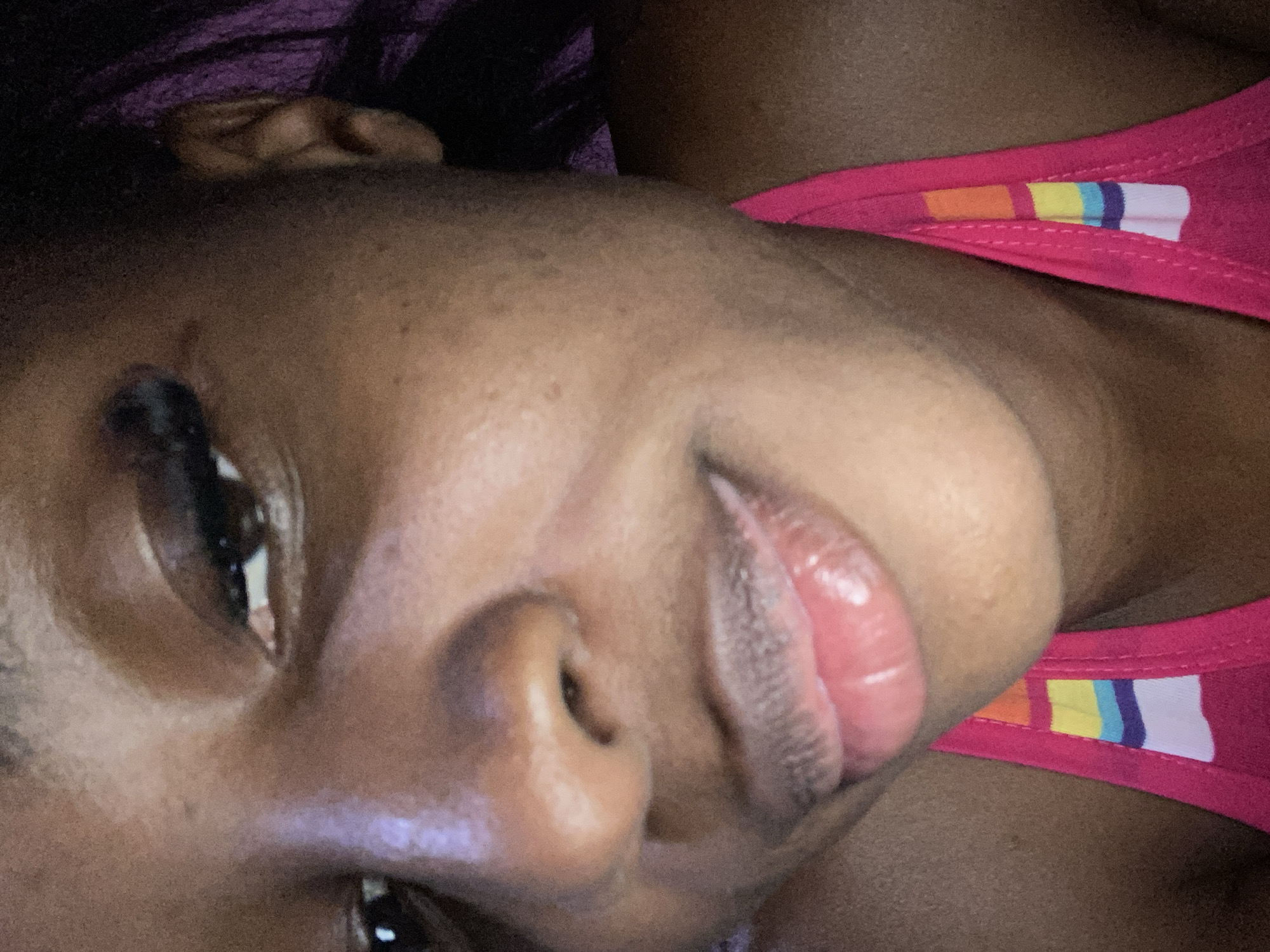 The image is a close-up, low-angle, indoor, selfie-style photograph of a Black person's face and upper chest. The person's face is angled slightly upwards and to their left, with their right eye more prominently visible. The skin tone is dark. The eye visible in the foreground is dark, with visible eyelashes and eyeliner. The person's lips are full and appear to have lip gloss. The nose is wide. The person is wearing a bright pink halter top with a rainbow-striped detail, consisting of horizontal blocks of orange, yellow, blue, and white. The background is dark and out of focus, with hints of purple in the upper right corner, possibly indicating hair or fabric. The lighting appears to be artificial and somewhat harsh, creating highlights on the skin. No specific location within Jos, Nigeria, can be identified from the image.