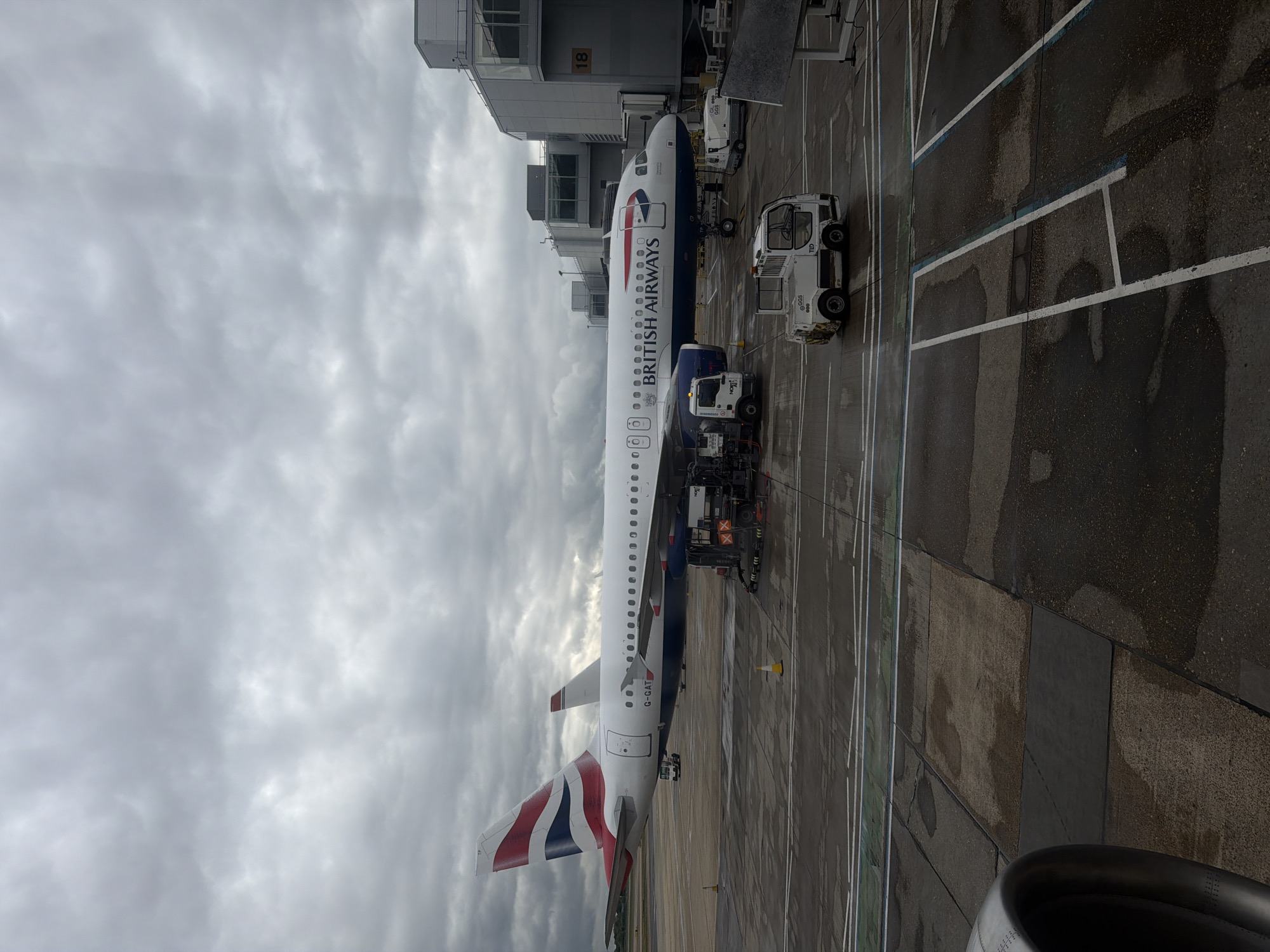 The image depicts a British Airways Airbus A320 series aircraft parked at an airport gate, likely at London Gatwick Airport, located in Crawley, United Kingdom. The aircraft is connected to ground service vehicles, including a refueling truck and a baggage towing tractor. The weather appears overcast with a cloudy sky. A portion of the airport terminal is visible adjacent to the aircraft's nose. Markings on the tarmac are visible including white taxiway lines. An engine nacelle is partially visible in the lower right corner of the image.