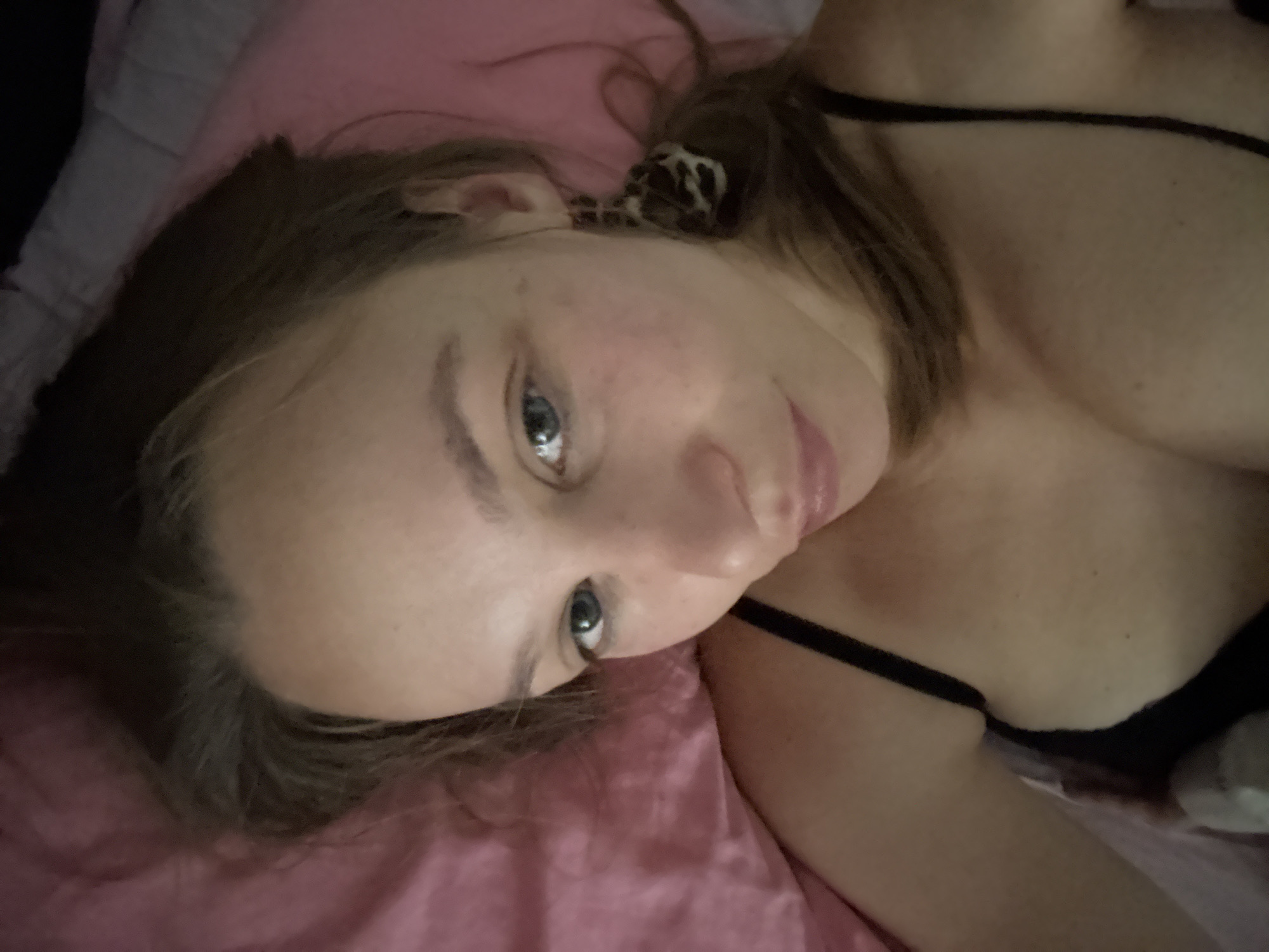 A woman with light brown hair and blue eyes is lying in bed, looking at the camera. She is wearing a black bra and a leopard-print earring. The bed is covered with a pink blanket. The scene appears to be in a bedroom. There is no information in the image to suggest the location in the city of Überherrn, Germany.