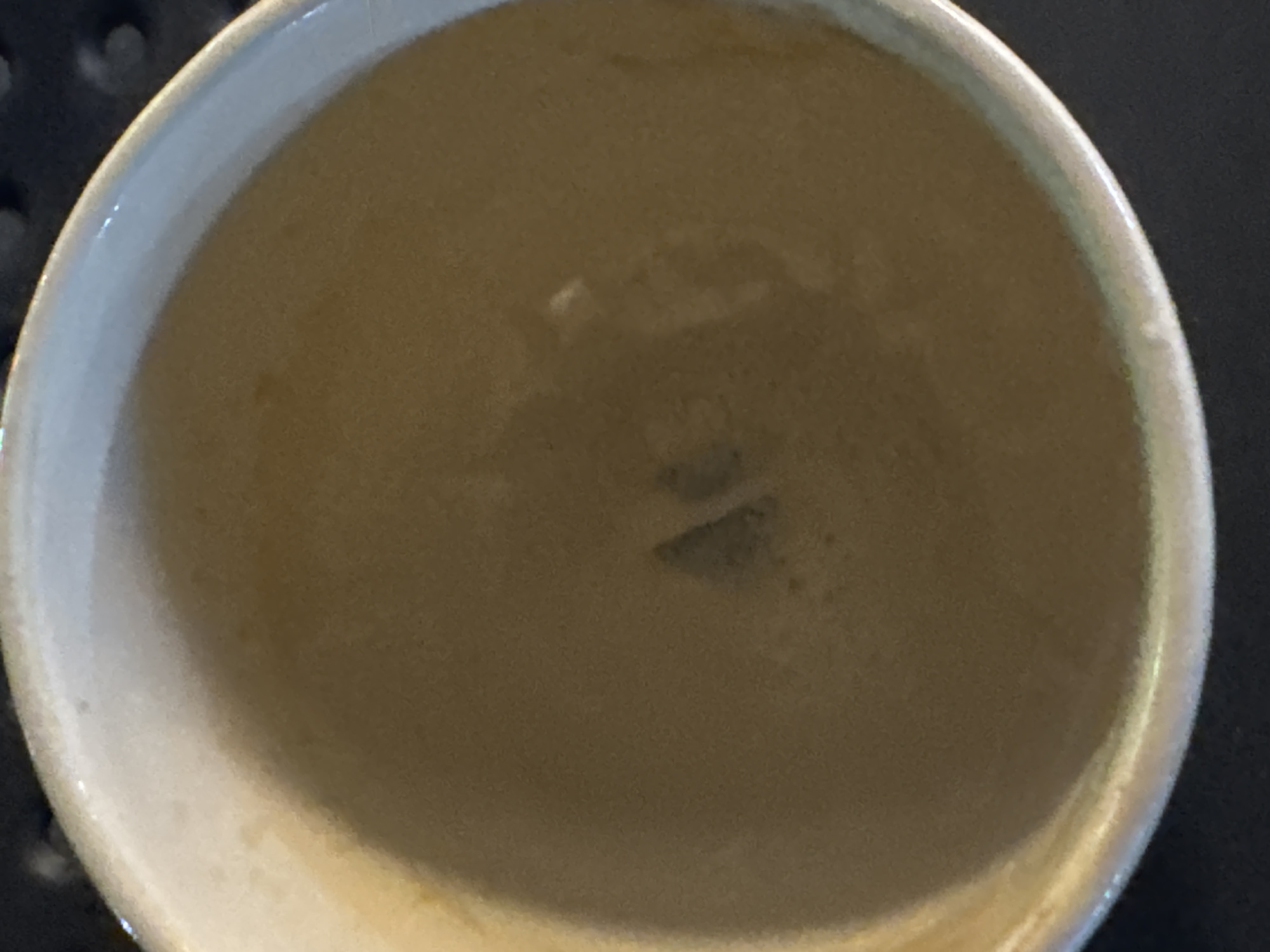 An overhead shot captures a close-up of a light-colored mug filled with a frothy, light brown liquid, likely coffee. The mug's rim is visible, appearing slightly chipped in places, with a subtle sheen suggesting it might be ceramic. The liquid inside has a creamy texture, with a small, darker, triangular-shaped object partially submerged in the center. The background is dark and out of focus, hinting at a table or countertop. No people or specific landmarks of Batumi, Georgia, are identifiable in this image.