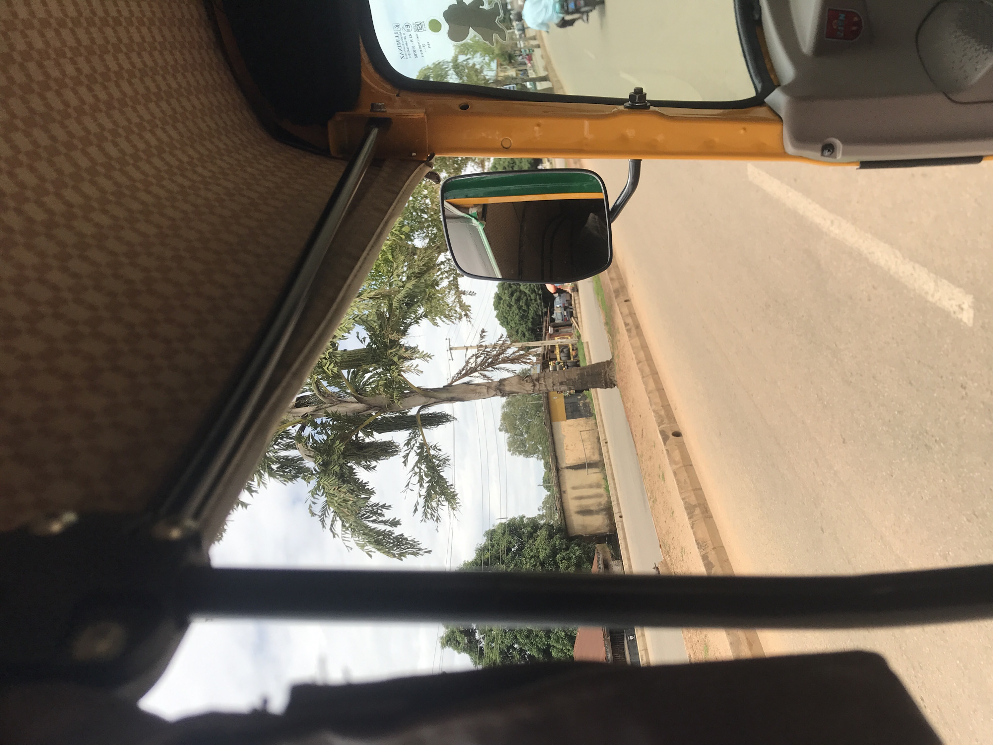 This image, captured from the interior of a yellow three-wheeled rickshaw, depicts a street scene in New Stadium, Nigeria, under an overcast sky. The vehicle's beige, checkered ceiling is visible in the upper portion, along with a dark horizontal support bar. On the right, a black side-view mirror with a green outer rim reflects the yellow interior of the vehicle and a green structure, while a dark silhouette sticker is affixed to the adjacent windshield. A blurry, dark vertical support column from the vehicle's frame partially obstructs the left side of the view.
Outside, a light brown paved road with a faint white line on its right edge extends into the distance, bordered by a concrete curb and a dusty shoulder. Several palm trees with green fronds and other leafy green trees line the roadside. Beyond the vegetation, single-story buildings with weathered, light-colored walls (some with yellow sections) and dark roofs are visible. Utility poles with overhead wires crisscross against the cloudy sky. In the mid-ground, a person wearing a light blue shirt is riding a motorcycle away from the viewer on the road, with other indistinct figures and structures present in the distant background.