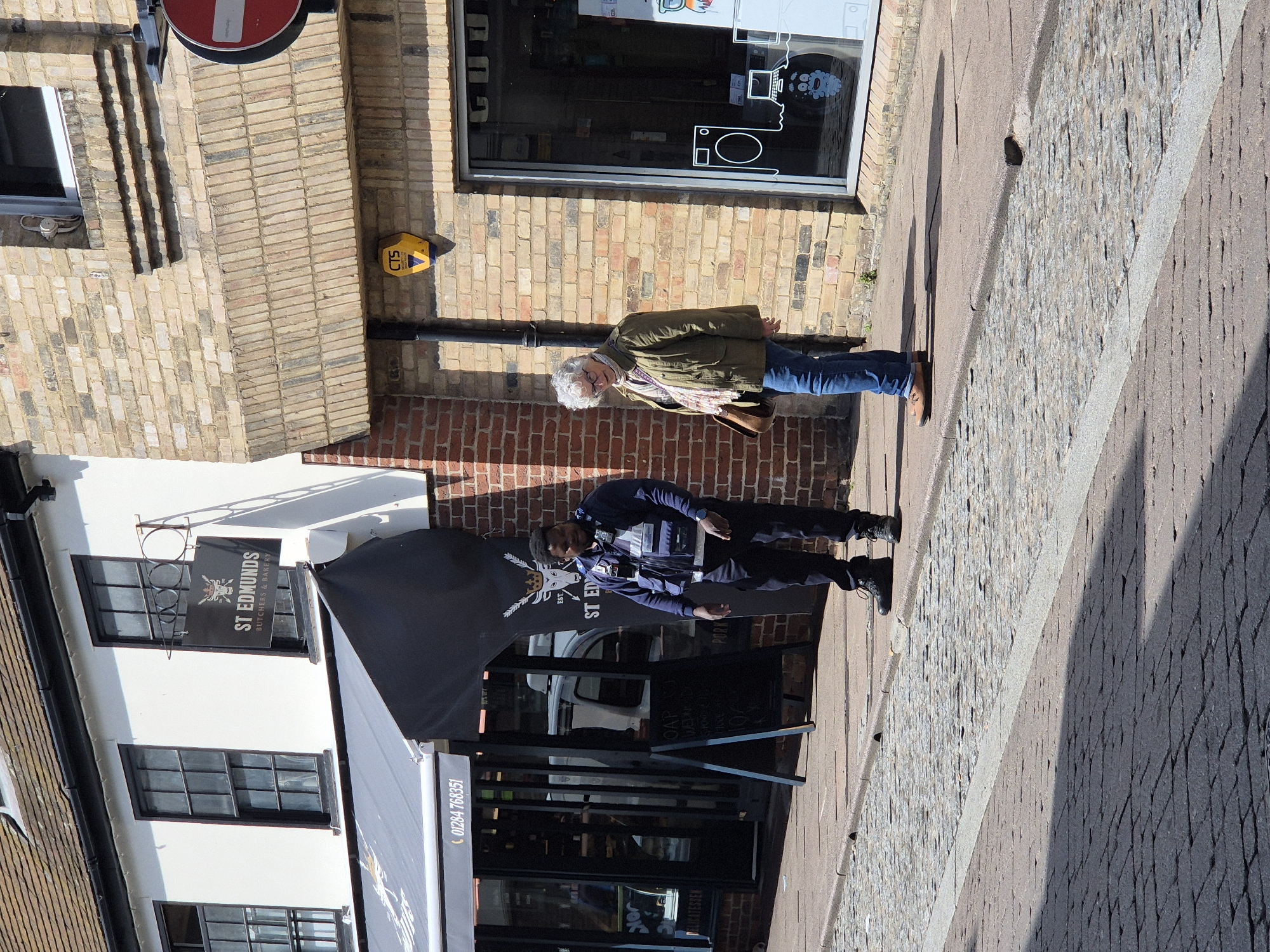 The image shows an outdoor scene in the city of West Suffolk, United Kingdom. In the foreground is a sidewalk paved with cobblestones. An awning is on the left side with "ST EDMUNDS BUTCHERS & BAKERY" written on it. The business phone number is "01284 768351". There are two people standing in front of the storefront. One is wearing a jacket and the other a blue uniform. A building with brick walls and windows can be seen in the background. A yellow "CTS" sign is fixed on the wall. There is also a "no entry" sign present. There's text "OAP" and "PORK" written as well.