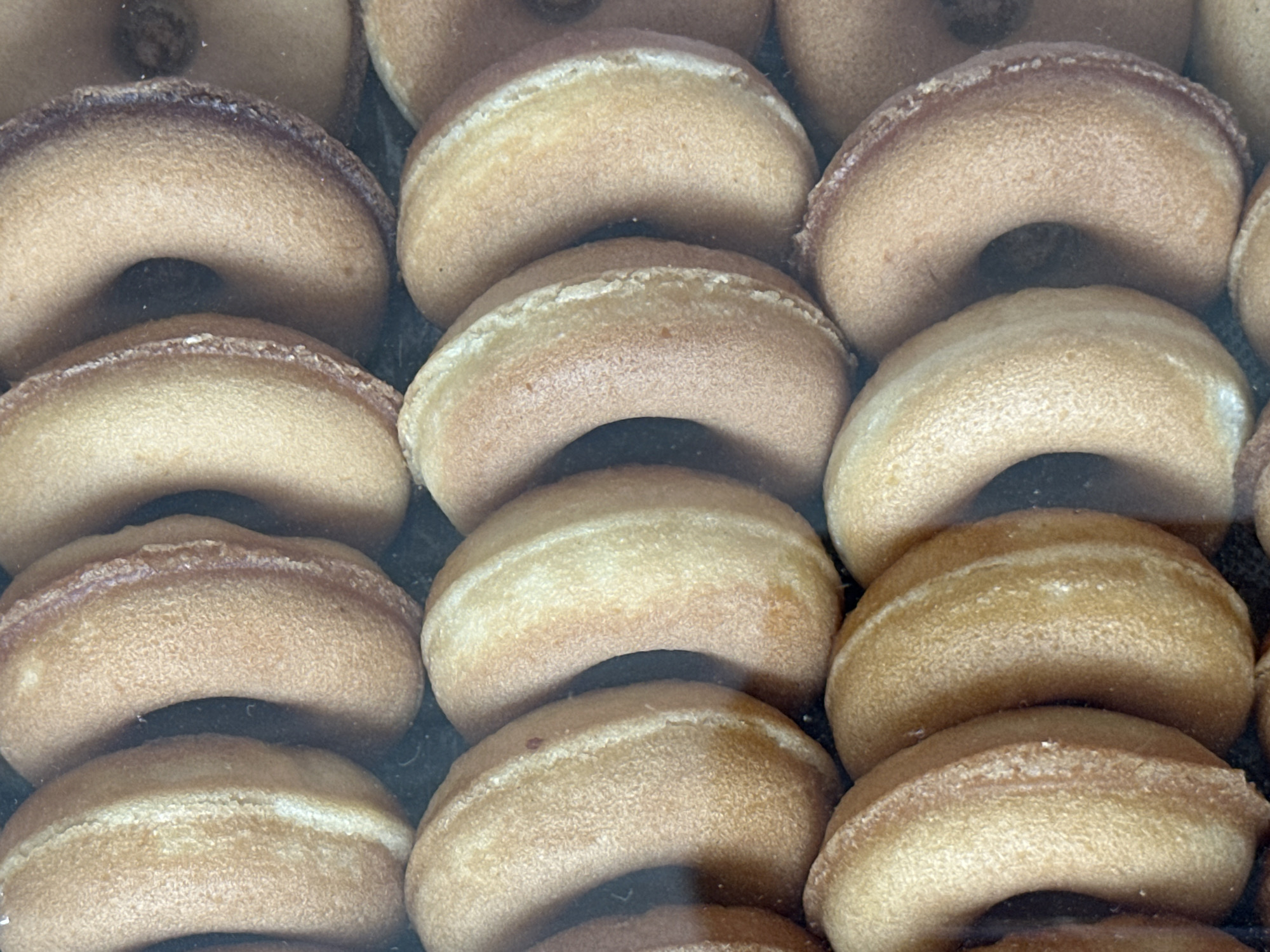 The image displays a close-up, top-down view of a tray of plain, ring-shaped donuts. The donuts are arranged in rows and columns, filling the frame. They appear to be baked, with a light brown, slightly textured surface. The lighting is even, highlighting the uniform color and shape of the donuts. The background is dark and out of focus, suggesting the donuts are inside a display case or packaging. There are no people or other objects visible. The location in Özgön, Kyrgyzstan, is not discernible from the image itself.