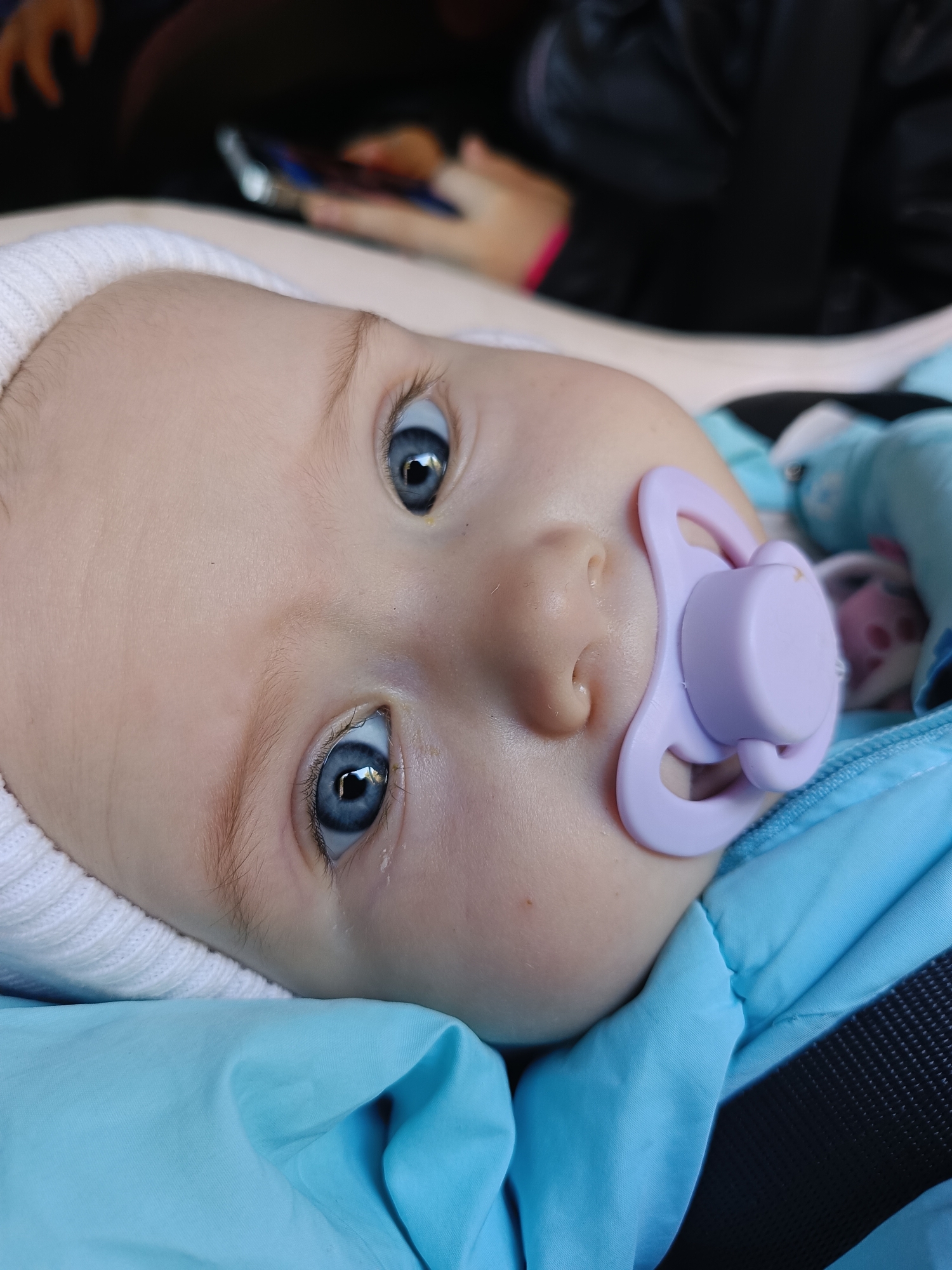The image is a close-up portrait of a baby, oriented with its head tilted to the left and looking directly towards the viewer. The baby has fair skin, delicate features, and bright, wide blue eyes that stand out prominently. Fine, light hair is visible beneath a white, ribbed knit hat that covers the baby's head. A light purple pacifier is comfortably in the baby's mouth. A small tear is visible near the inner corner of the baby's left eye, though the overall expression appears calm and curious.
The baby is nestled in or dressed in vibrant blue quilted material, which could be part of a warm jacket, snowsuit, or a cozy blanket. On the right side of the blue fabric, a black, textured strap, consistent with a car seat or stroller harness, is partially visible, suggesting the baby is secured in a carrier. The background is softly blurred, showing a dark environment where the vague outlines of an adult's hand, holding a mobile phone, can be discerned, indicating the presence of another person. A light-colored, indistinct structure is also present in the upper left background.
The scene appears to be set indoors or within a vehicle. No specific time of day or weather conditions can be determined from the image itself. The provided context indicates the location is Chaikovsky, Russia. There is no visible text within the media file.