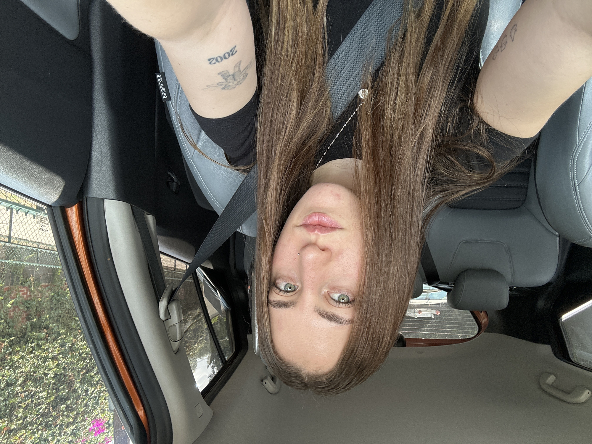 The image, originally oriented upside down, depicts a young person, likely female, seated inside a car in Mexico City during the daytime. The individual has long, straight brown hair, light skin, and striking green/hazel eyes, looking directly into the camera with a neutral expression. A philtrum piercing is visible above her upper lip. She is wearing a black long-sleeved top and a grey seatbelt across her chest.
Visible tattoos include the year "2002" with a bird or swallow design on her right arm and three small, abstract shapes on her left arm. The car's interior features light grey leather or vinyl seats. A prominent "SRS AIRBAG" label is visible on the seatbelt pillar near her right shoulder.
Through the car windows, lush green foliage and a chain-link fence are visible, suggesting an urban or suburban setting. An orange-colored trim is visible along the car's door frame. In the rearview mirror, part of another vehicle, possibly a truck or van, can be seen. Reflections on the window also show reversed text, including "2AZFE." The bright lighting indicates it is likely a clear, sunny day. The overall scene captures a moment inside a parked or stationary car.