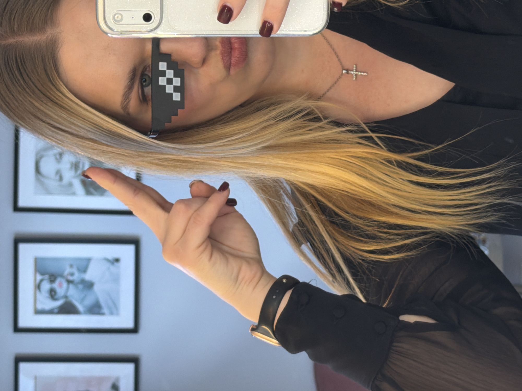 A woman with blonde hair and "deal with it" pixel sunglasses is taking a selfie. She is wearing a black blouse and a necklace with a cross pendant. She's holding a phone with maroon nail polish. Behind her are framed pictures on a light blue wall. The location is unknown and there is no information to confirm the location is Riga, Latvia.