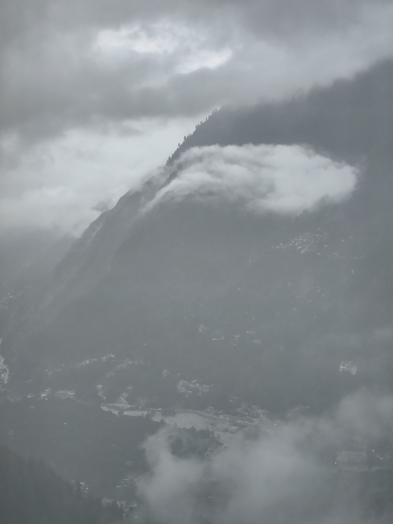 This image depicts a misty mountain landscape in Prini, India, with a village nestled in the valley below. The sky is overcast with heavy clouds, contributing to the diffused light and muted color palette. The foreground is dominated by the imposing presence of a mountain range, its slopes partially obscured by swirling mist and low-hanging clouds. A patch of dense foliage, likely pine trees, can be seen near the crest of one of the mountains. In the valley, the village of Prini is visible, a cluster of structures with faint lights suggesting human habitation. The misty conditions create a sense of depth and atmosphere, blurring the details of the distant landscape and lending an ethereal quality to the scene. The overall impression is one of a serene, albeit somber, mountain environment under a cloudy sky.