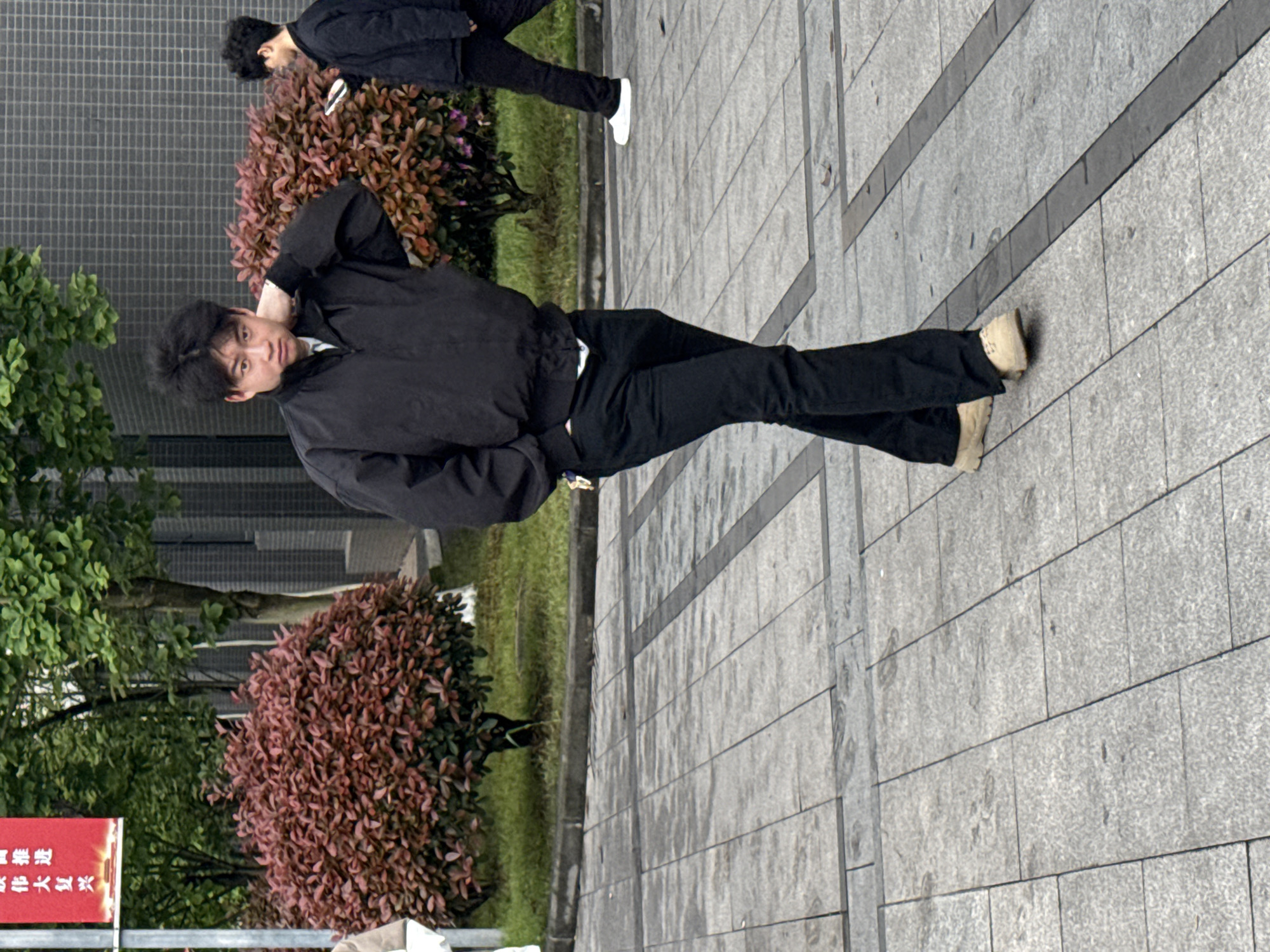 The image shows a person, possibly male, standing on a paved sidewalk in an urban setting. The person is wearing dark clothing, including a jacket and pants, and light-colored shoes. He is looking directly at the camera with his left hand raised to his neck. Behind him are shrubs and a wall with a grid pattern. In the background, another person is walking away. A red sign with Chinese characters is visible in the lower left corner, partially obscured by foliage. The pavement is gray with darker gray lines running across it. The photo appears to be taken during the day, with natural lighting. Based on the sign with Chinese characters, the picture is most likely taken in China, possibly in Jiangjin.