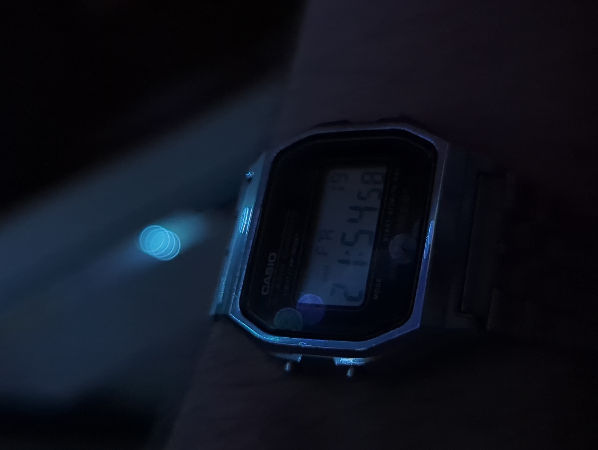 A close-up, low-light shot of a Casio digital watch on a person's wrist, displaying the time 21:54:58 and the day FR (Friday) 19. The watch is silver-toned with a black face and digital display. Blue light sources create visible lens flare and bokeh effects in the upper portion of the image. The background is dark and indistinct, suggesting an indoor setting at night or in a dimly lit environment. No specific location in Votkinsk, Russia, can be identified from the provided image.