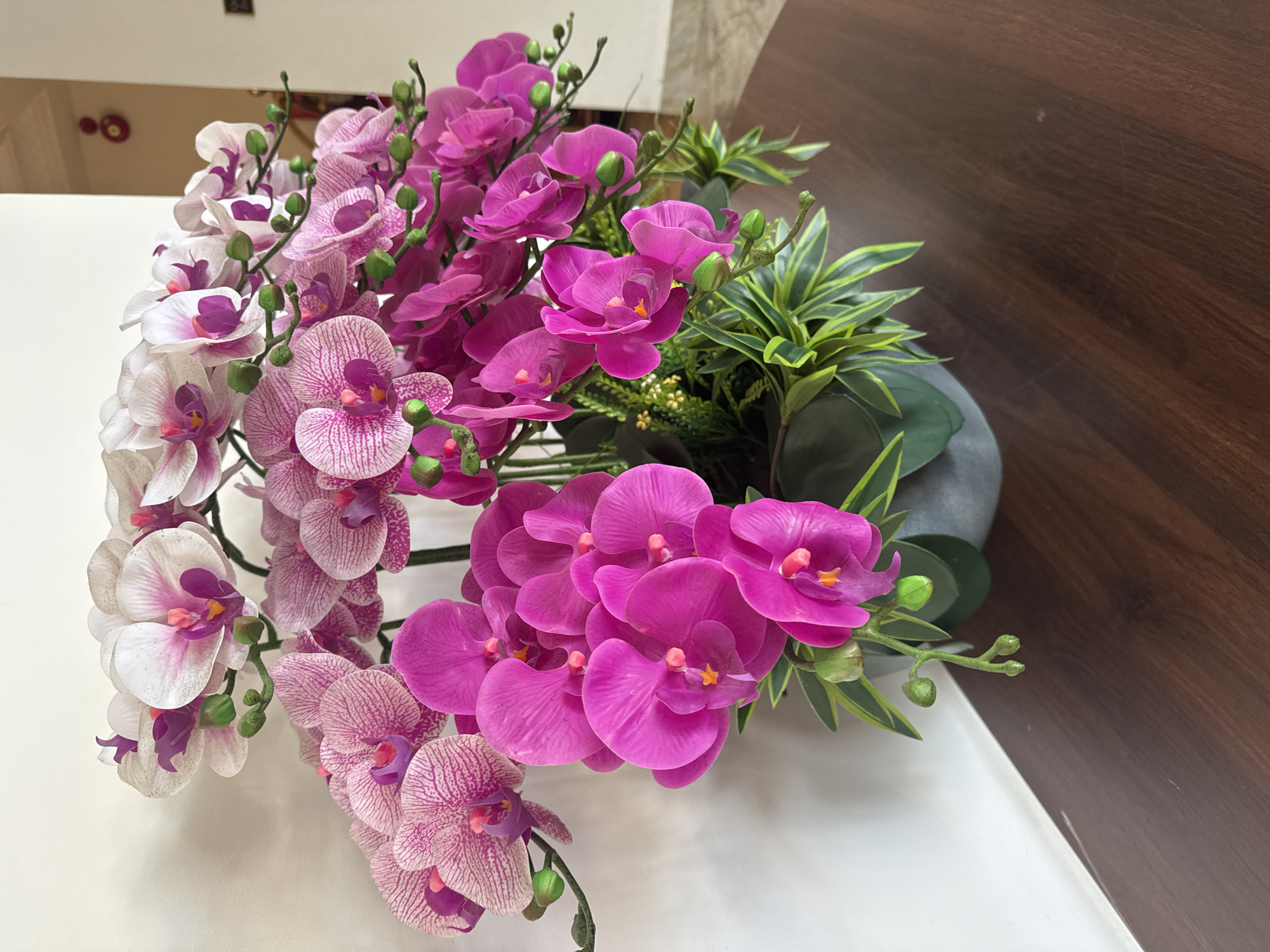 The image depicts a floral arrangement featuring artificial orchids in shades of white, pink, and magenta, alongside green foliage. The flowers are arranged in a gray container. The backdrop consists of a white surface and a dark wood panel. The location is not discernible from the image. No people, specific actions, or interactions are visible.