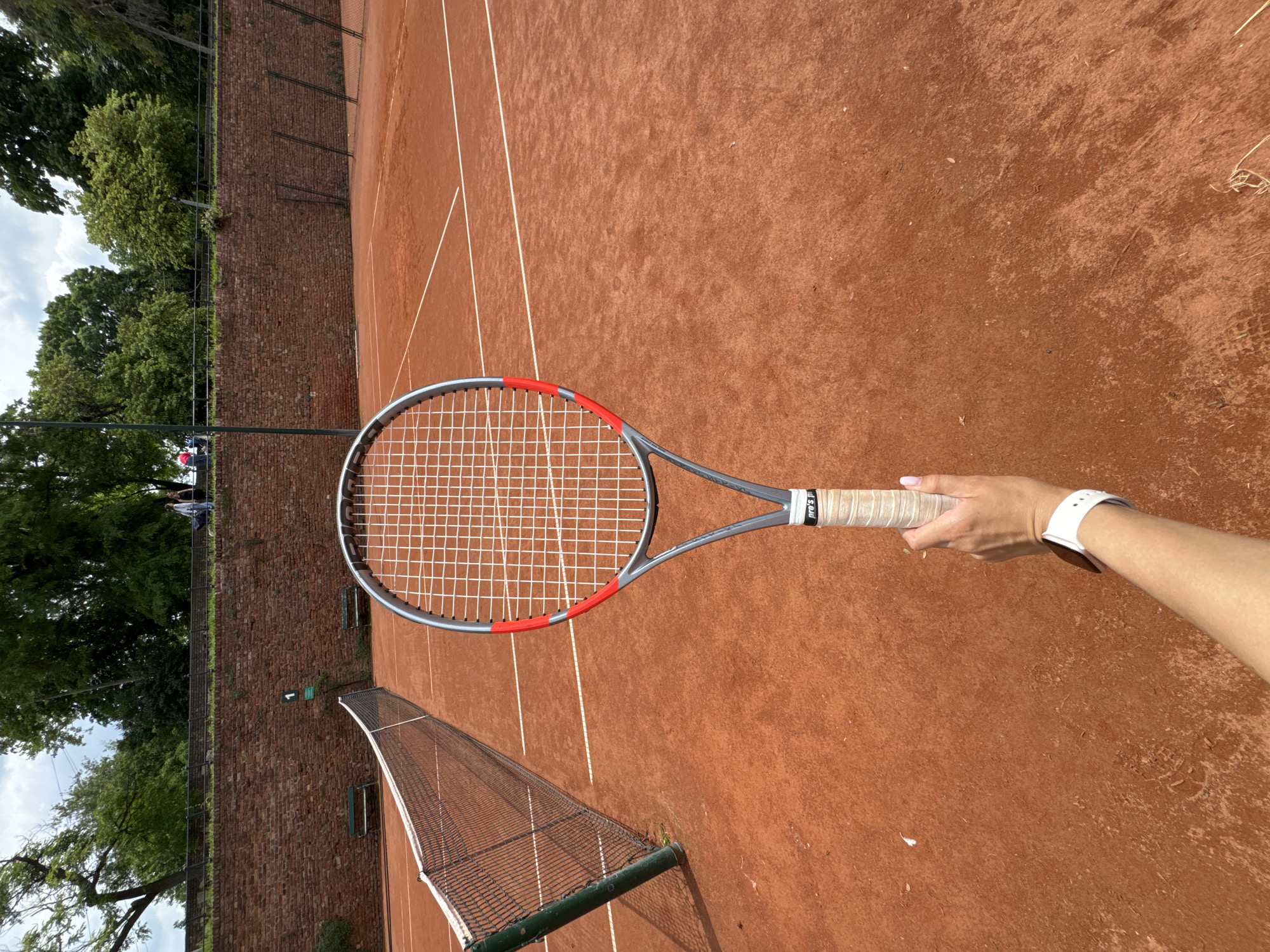 The image shows a tennis court in Belgrade, Serbia. The court is made of red clay and has white lines marking the boundaries. A tennis net divides the court. The court is surrounded by a brick wall and trees. A person holds a tennis racket in their hand. The person wears a white watch. The background includes a glimpse of people on a tennis court.