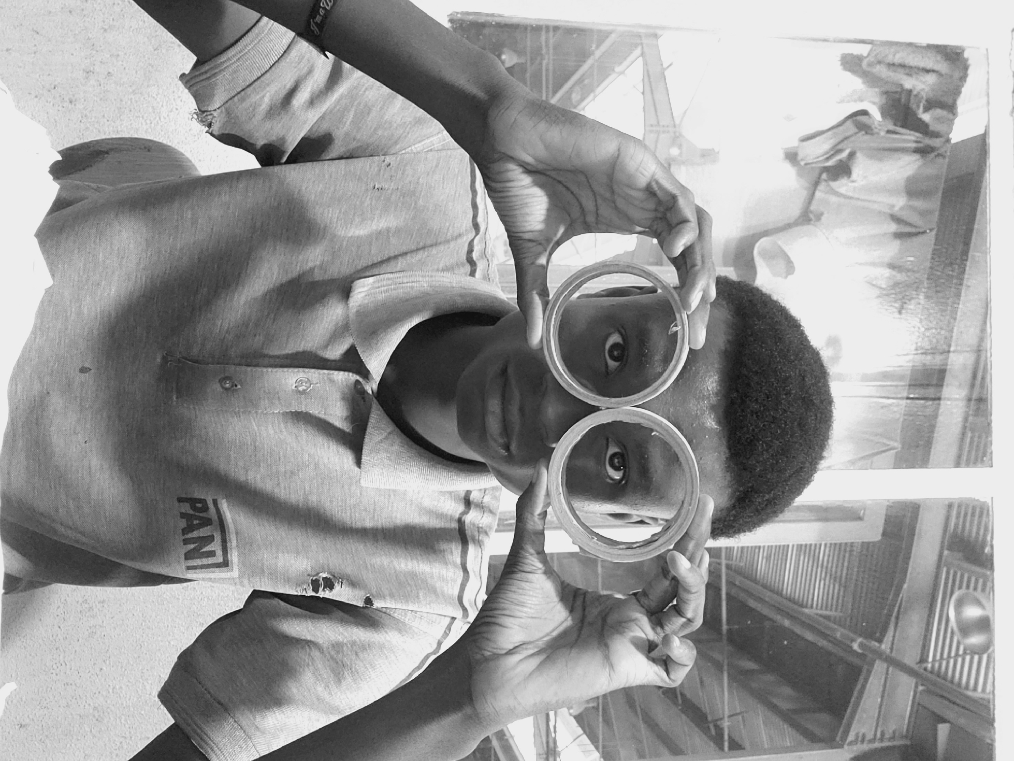 A black and white, vertically oriented photograph depicts a young person, likely male, from an African ethnicity, looking directly at the viewer. The subject is positioned roughly waist-up, seen from a slightly elevated angle. They wear a light-colored, short-sleeved collared polo shirt with a striped design on the front and visible wear, including small holes. The text "PAN" is clearly visible on the lower left side of the shirt. A dark bracelet with partially visible text, possibly starting with "I'm a...", is worn on the right wrist.
The person holds two clear, circular objects, resembling plastic rings or lenses, up to their eyes like makeshift spectacles. Their dark, short curly hair is visible. The eyes are wide and focused, seen through the rings, creating a playful or inquisitive expression. The background appears to be a reflective surface, possibly a window or a sheet of metal, showing reflections of what looks like a corrugated metal roof or ceiling structure and possibly other elements of an indoor space, suggesting an industrial or utilitarian setting. The lighting is bright, indicating daytime. The scene captures a moment of engagement, potentially playful or curious, in Kakuri Gwari, Nigeria.