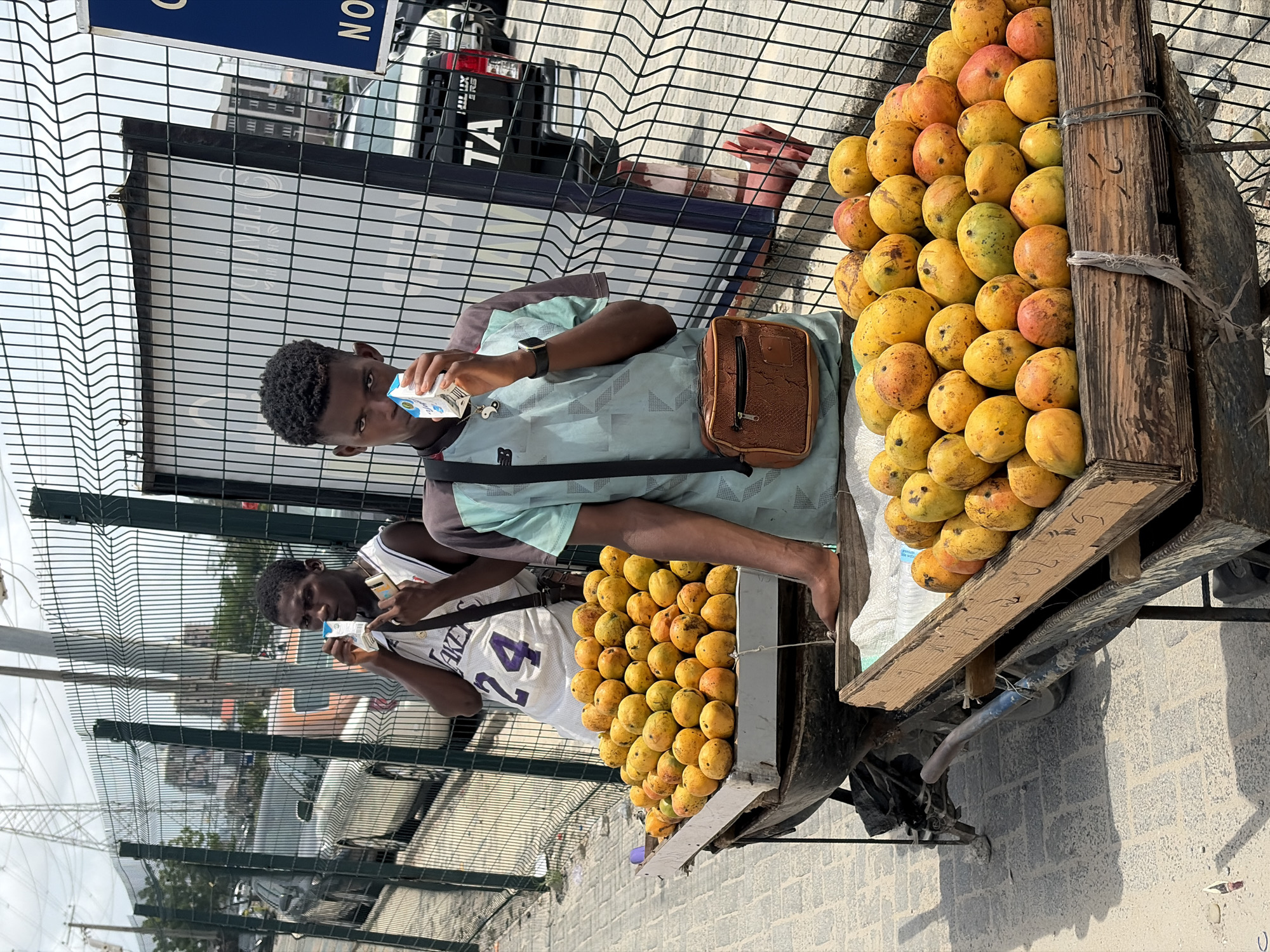 In Igboefon, Nigeria, two Black men stand near a fence and a cart laden with mangoes. The man closest to the camera wears a white Lakers jersey with the number 24. He's holding a small, white juice box and looking off-camera. The second man wears a light blue t-shirt and a brown crossbody bag. He also holds a juice box, drinking directly from the container. The cart is piled high with ripe, yellow mangoes, stacked neatly on a wooden platform. The fence is a dark grey, and a vehicle is visible behind it. The ground is paved, and the lighting suggests it's a sunny day.