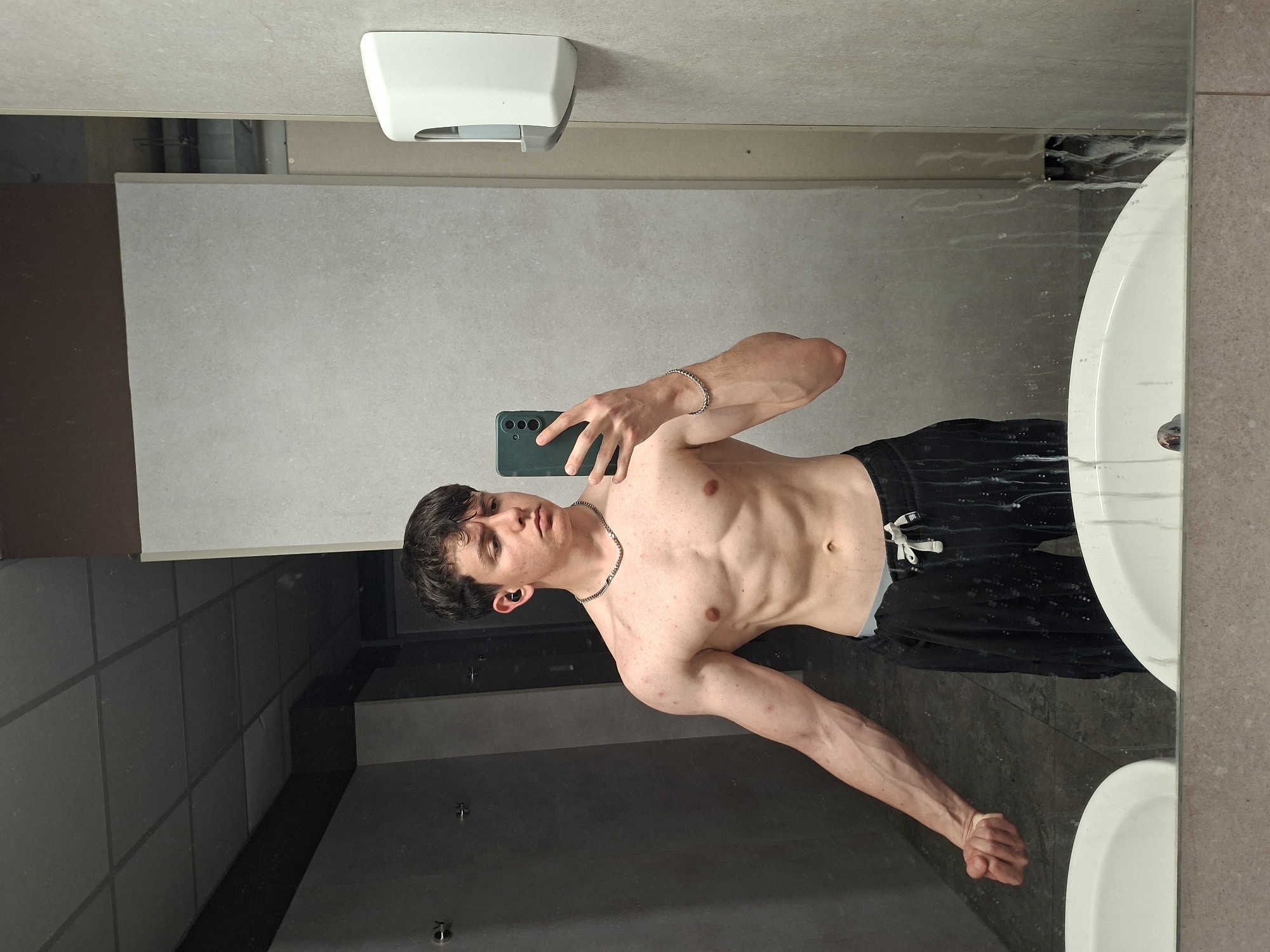 A young, shirtless man is taking a mirror selfie, showcasing his muscular physique with a toned abdomen and defined arms. He is wearing black athletic shorts with a white drawstring, a silver chain necklace, a silver bracelet on his right wrist, and a dark earbud in his left ear. He holds a dark green smartphone in his right hand, angled to capture his reflection. His left arm is extended downwards, highlighting the musculature. The setting is an indoor space, likely a public restroom or changing area, with light grey tiled walls. A white hand dryer or dispenser is mounted on the wall above his head. A portion of a white sink is visible in the reflection on the right side of the mirror, which also shows some smudges or water marks. The floor, reflected in the mirror, is dark-tiled with visible grid patterns. The man looks intently at the camera, posing confidently. The image depicts a casual moment of self-documentation in Plovdiv, Bulgaria. No visible text is discernible.