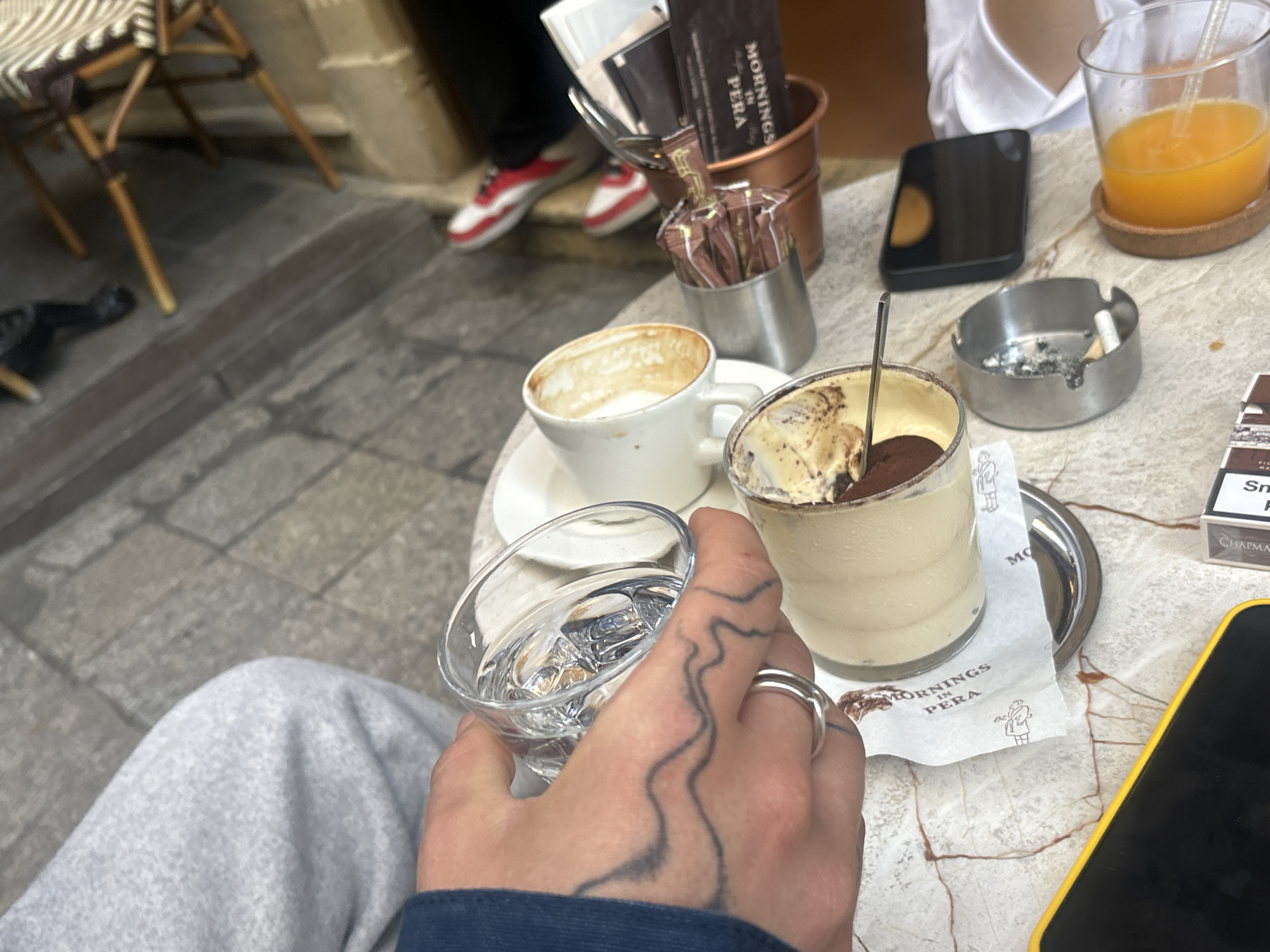 The image depicts an outdoor cafe scene in the Beyoğlu district of Istanbul, Turkey. A person is holding a glass of water with ice, their hand tattooed with a wavy line design and adorned with a silver ring. On the marble-patterned table are a partially consumed cup of coffee, a glass of tiramisu, an ashtray with a cigarette butt, a cell phone, a glass of orange juice, a packet of cigarettes, a small bucket of sugar packets labeled "Mornings in Pera", and an additional cup of various utensils. Other people are seated, but only their legs and sneakers are partially visible. A chair with a woven seat stands nearby on the stone pavement.