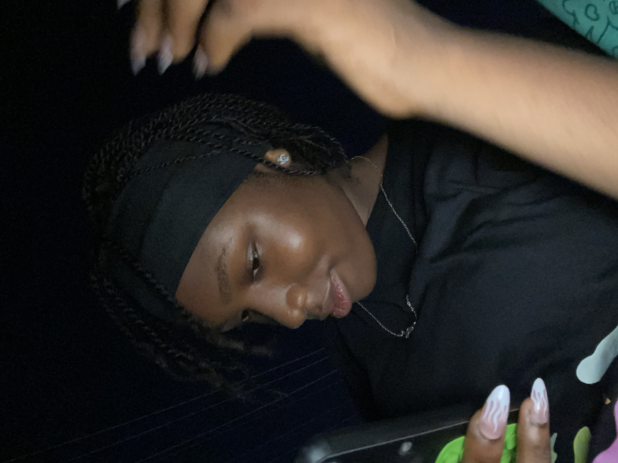 A person with dark skin and braided hair is pictured indoors, possibly in the city of Lagos, Nigeria, though the specific location isn't evident from the image itself. The person is wearing a black headband and a dark shirt. A silver necklace is visible around their neck, and they appear to be looking down at a mobile phone. Another person's arm and hand are visible at the top and bottom edges of the frame. The bottom arm displays an elaborate nail design with white and swirling accents. The lighting is subdued.