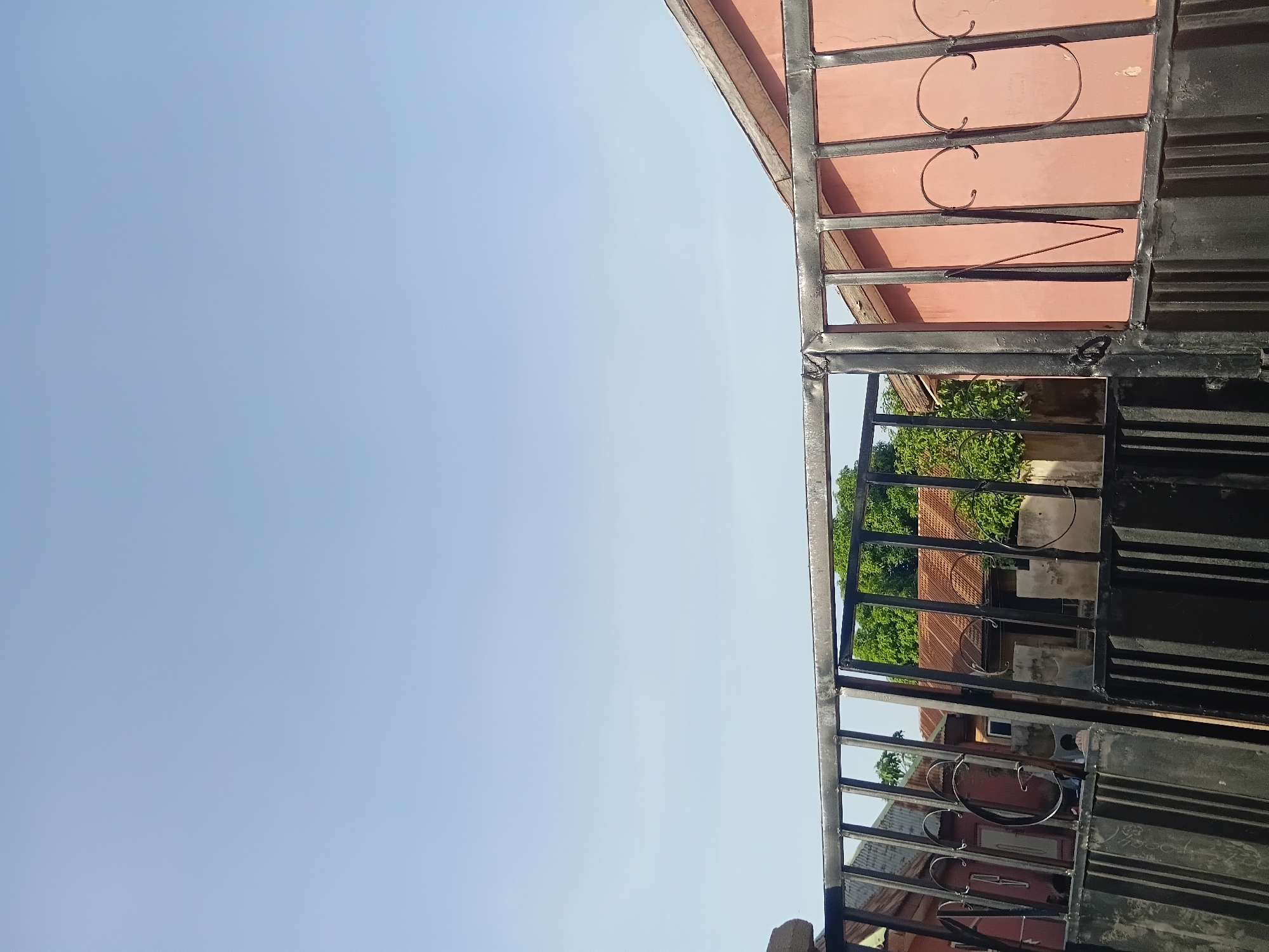 A wide-angle, eye-level shot captures a black metal gate in the foreground, partially obscuring a glimpse of buildings with red and brown roofs and lush green trees behind it. The sky is a clear, pale blue. The gate appears to be a residential entrance, with some decorative swirls on the right side. The buildings suggest a typical urban or suburban setting in Jalingo, Nigeria. No people are visible in the frame. The lighting indicates it is daytime, likely morning or late afternoon due to the soft light.
