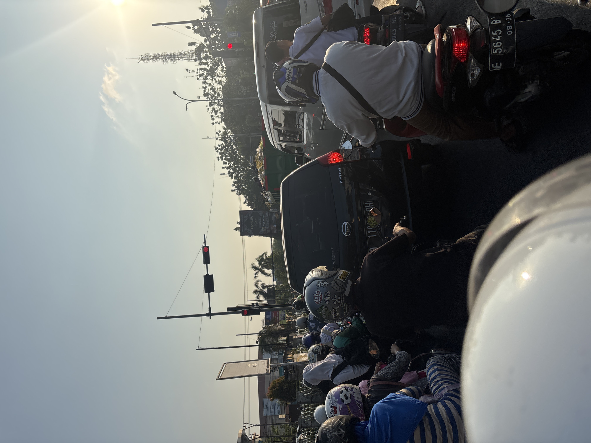 The image depicts a traffic jam on a city street in Cirebon, Indonesia, with numerous motorcycles and a few cars. The sky is clear and bright, with the sun shining, indicating it is daytime. The traffic light is red, causing vehicles to halt. The motorcycles are densely packed, with several riders wearing helmets. People are dressed in casual attire, including one individual in a blue and striped shirt and another in a white shirt. In the background, buildings and trees are visible, along with a telecommunications tower. The overall scene conveys a typical urban traffic situation.