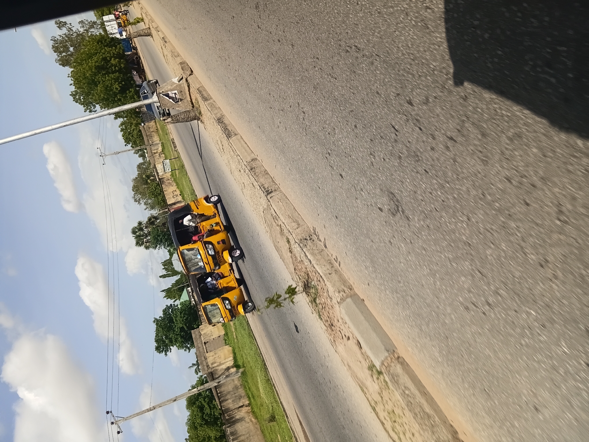 Two yellow auto rickshaws, also known as Keke Napep, are traveling on a paved road in Jalingo, Nigeria. The auto rickshaws are a common mode of public transportation in Nigeria, and they are often used to transport passengers and goods in urban areas. The road is a two-lane road with a curb and a sidewalk. The sidewalk is lined with trees and bushes. In the background, there are buildings and a wall. The sky is blue with some clouds. The scene appears to be a typical day in Jalingo, with the auto rickshaws moving along the road.