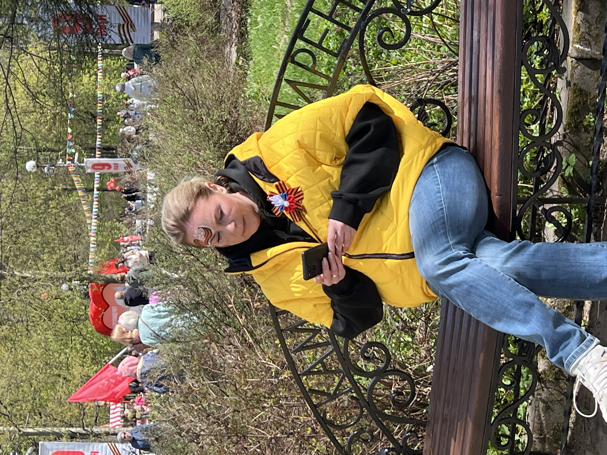 The image depicts a woman sitting on a park bench in Cherepovets, Russia. She is wearing jeans, a black hoodie, and a bright yellow quilted jacket. On her jacket, she has a ribbon in the colors of the Russian flag and the Saint George ribbon. She has a design painted on her face and is looking at a smartphone in her hands.
In the background, there is a crowd of people gathered, and there are several red flags and balloons visible, suggesting some kind of public celebration or event. A sign with "May 9" on it is visible in the background near the flags. The trees are in full foliage, indicating it is likely springtime. The bench is made of wood and wrought iron, with decorative elements.