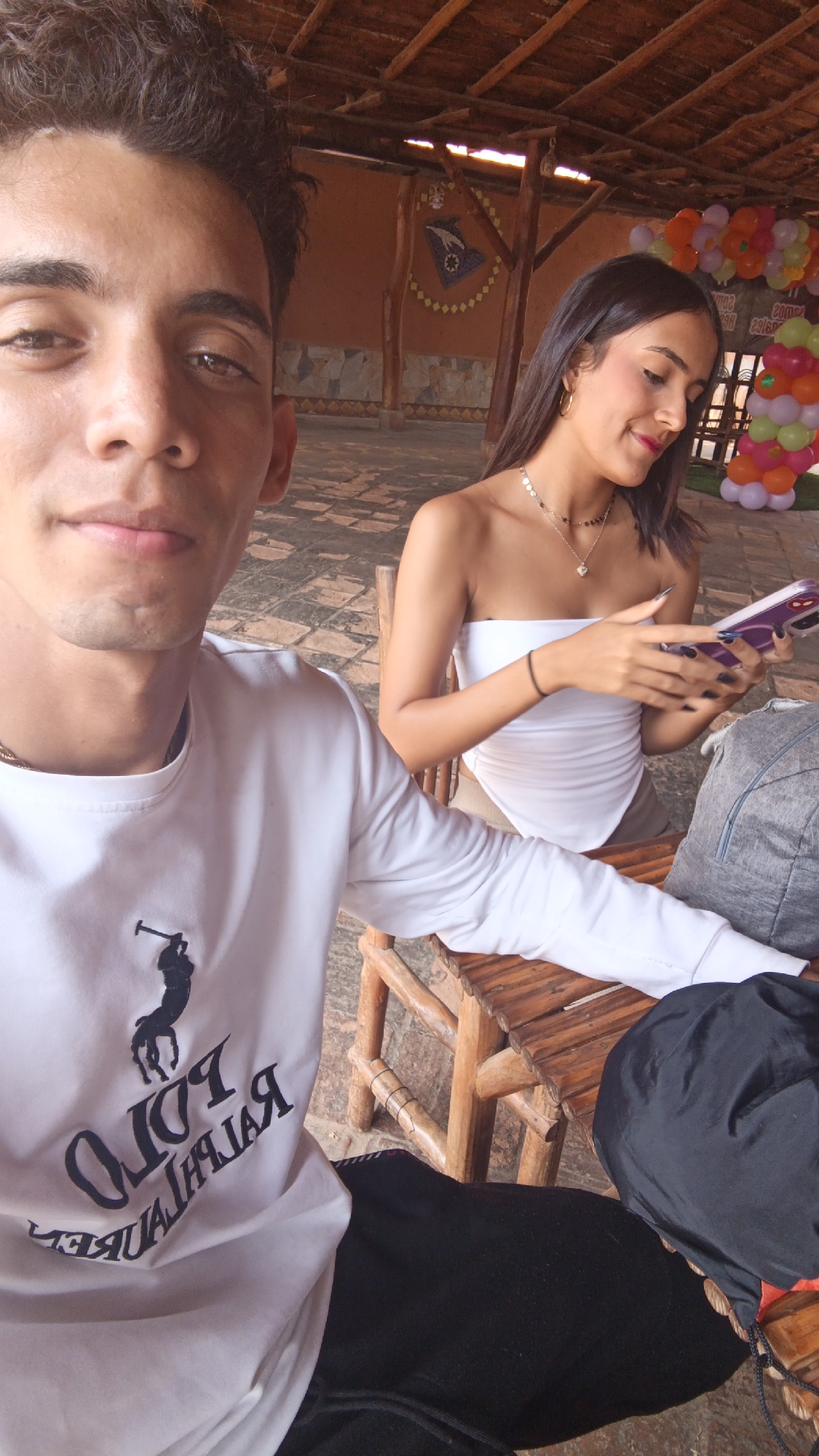 A selfie-style photograph depicts a young man and woman seated outdoors. The man, positioned in the foreground, has short, dark hair and is wearing a white long-sleeved shirt with a prominent Ralph Lauren logo. The woman, in the background, has long, dark hair and is wearing a white strapless top. She is looking down at a cell phone she holds in her hands. They are sitting at a wooden table with matching wooden chairs.
Behind them, there is a building structure with a thatched roof. A decorative emblem is visible on the wall, along with balloons in the background, suggesting a celebration. The flooring appears to be tiled.
This photo was taken in Juan Griego, Venezuela.