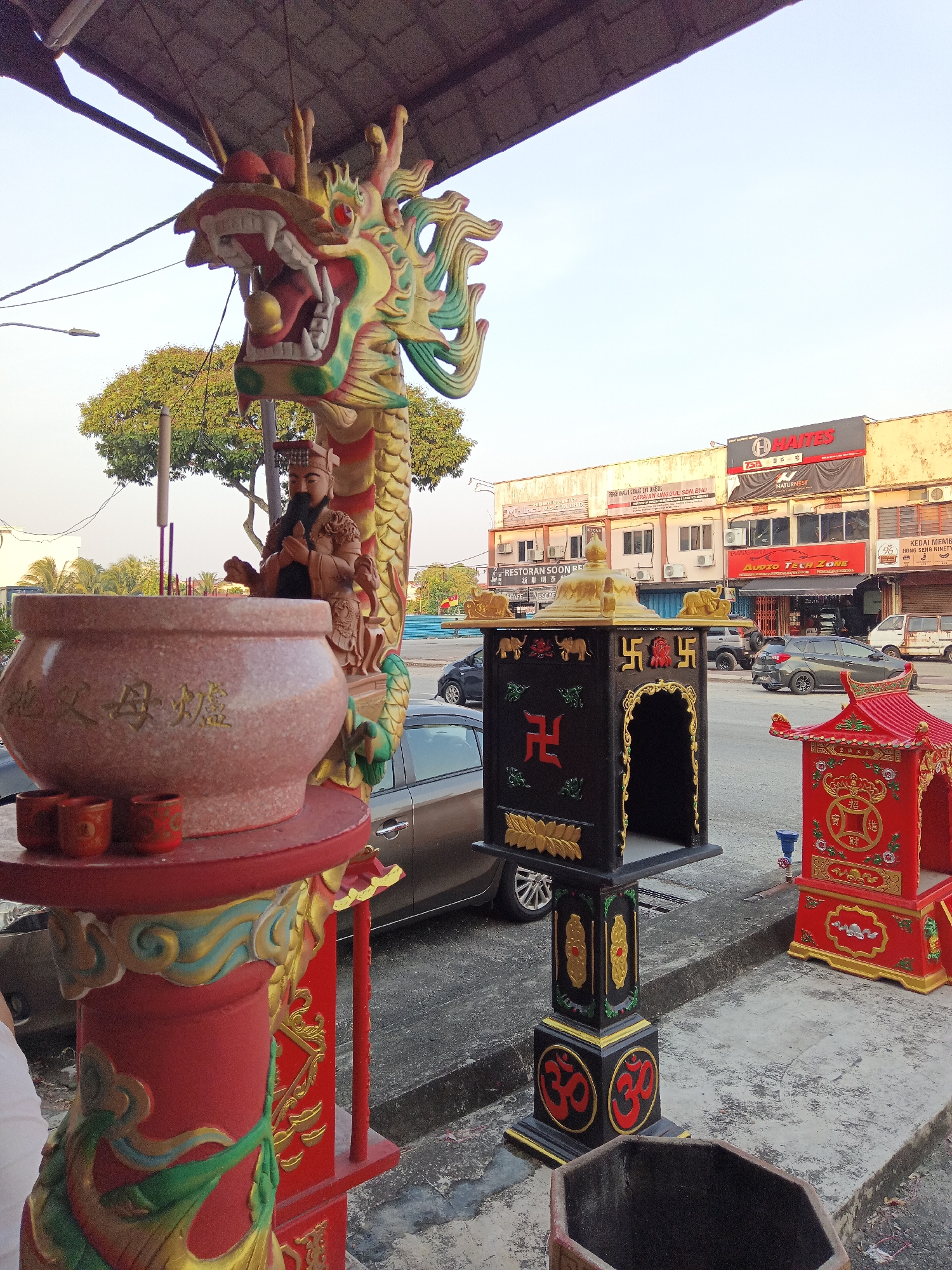 The image depicts an outdoor scene in Klang City, Malaysia, likely in front of a row of commercial establishments. In the foreground, a large pink incense burner with Chinese characters is prominently displayed on a red and gold ornate pedestal. The pedestal is adorned with a large, colorful dragon sculpture coiled around it. To the right of the incense burner are two smaller red cups. Further to the right, a black and gold altar-like structure stands, featuring Swastika symbols and elephant figures. Next to it, a smaller, bright red shrine-like object with gold detailing is visible. In the background, trees line the street, and a row of shops with various signage, including "HAITES," "NATURENEST," and "AUDIO TECH ZONE," are visible. Several vehicles are parked on the asphalt road and sidewalk. The sky is clear and bright, suggesting daytime. No people are clearly visible in the image, but a portion of a white garment is visible on the left edge, potentially belonging to someone holding the camera.