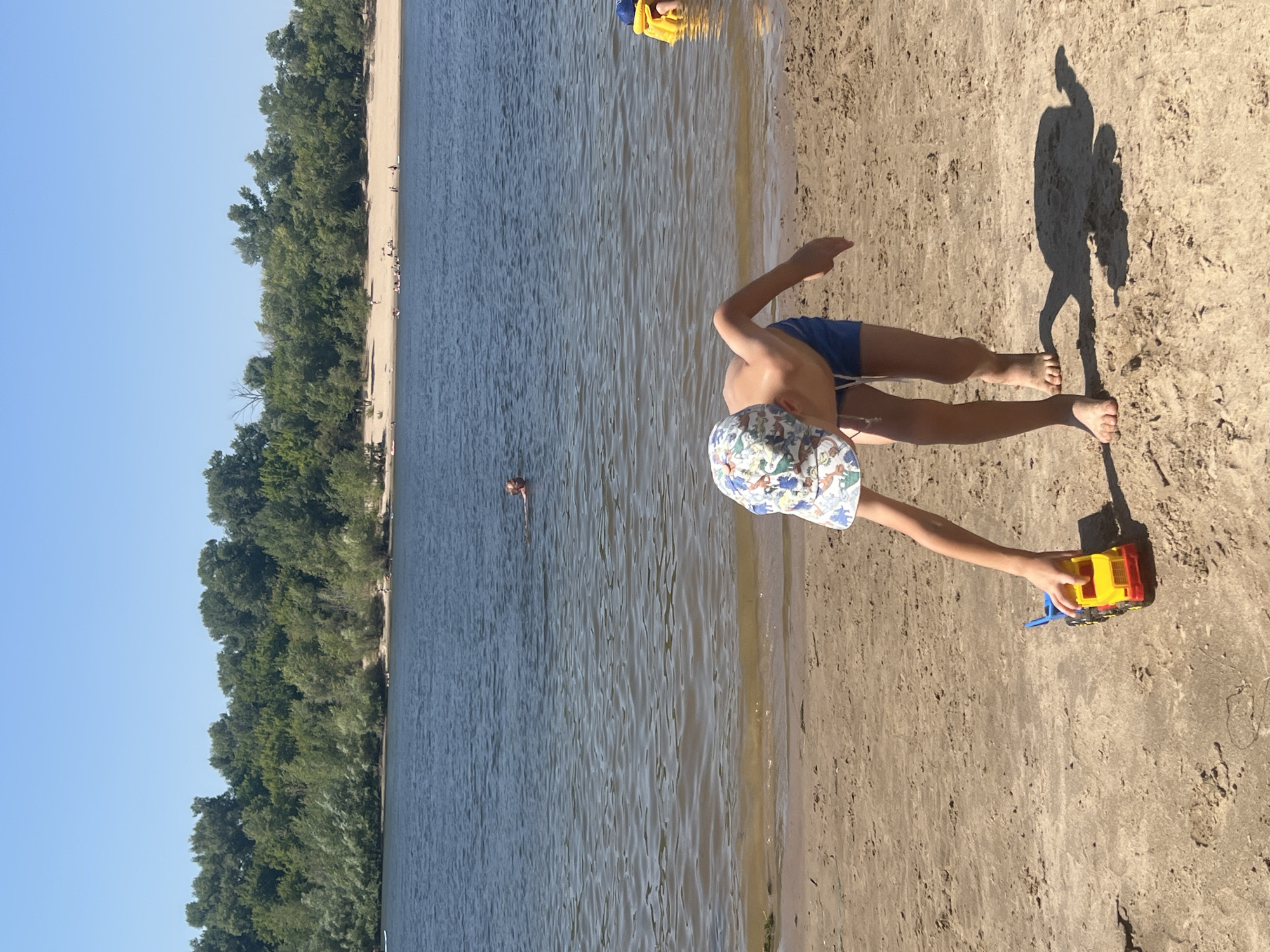 A young boy wearing a patterned hat and blue swim shorts is bent over on a sandy beach, reaching for a toy truck. His shadow is cast behind him on the sand. The beach is next to a body of water, likely a river, with a lush green treeline in the background. Several other people are visible in the water and on the distant beach, some swimming and others relaxing. The sky is clear and blue. This scene is located in Kilyakovka, Russia.