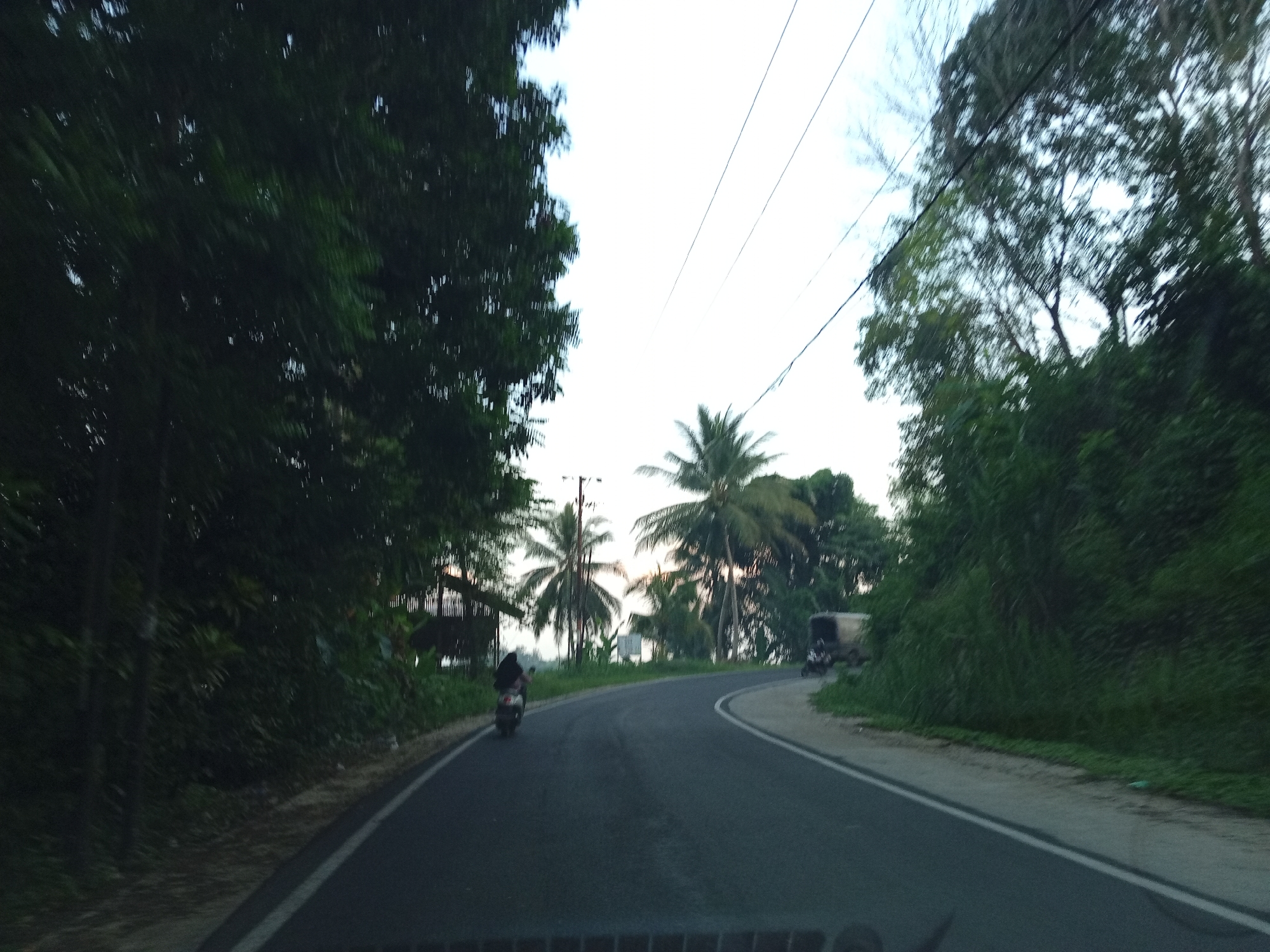 This is a photograph taken from a moving vehicle on a paved road, likely in Uleejalan, Indonesia. The road curves to the right, bordered by dense vegetation and trees on both sides. On the left side, a person is riding a scooter away from the camera. Further ahead on the road, another vehicle, possibly a truck, is visible in the distance. Several coconut trees are interspersed with other foliage. Utility poles and wires are present in the background. The sky is overcast, suggesting it might be dusk or a cloudy day. The overall scene depicts a rural or semi-rural road in a tropical environment.