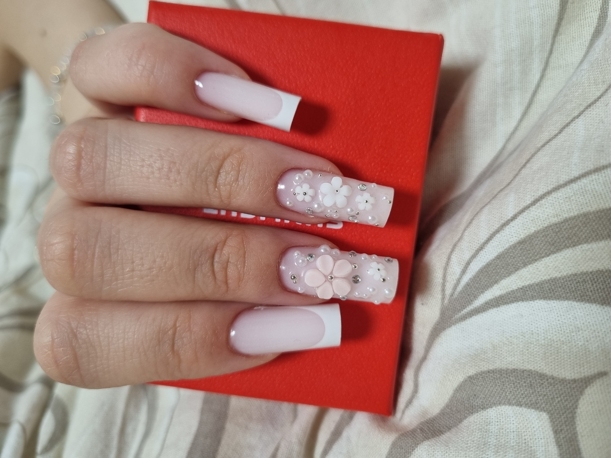 A close-up shows a hand holding a red, rectangular object. The hand has long, square-shaped fingernails with two nails having a white French manicure tip. One nail is decorated with small white flowers and silver rhinestones. Another nail is decorated with larger, light pink flowers and pearls. The background features a light-colored, striped surface. No location or people are explicitly identifiable in the image.