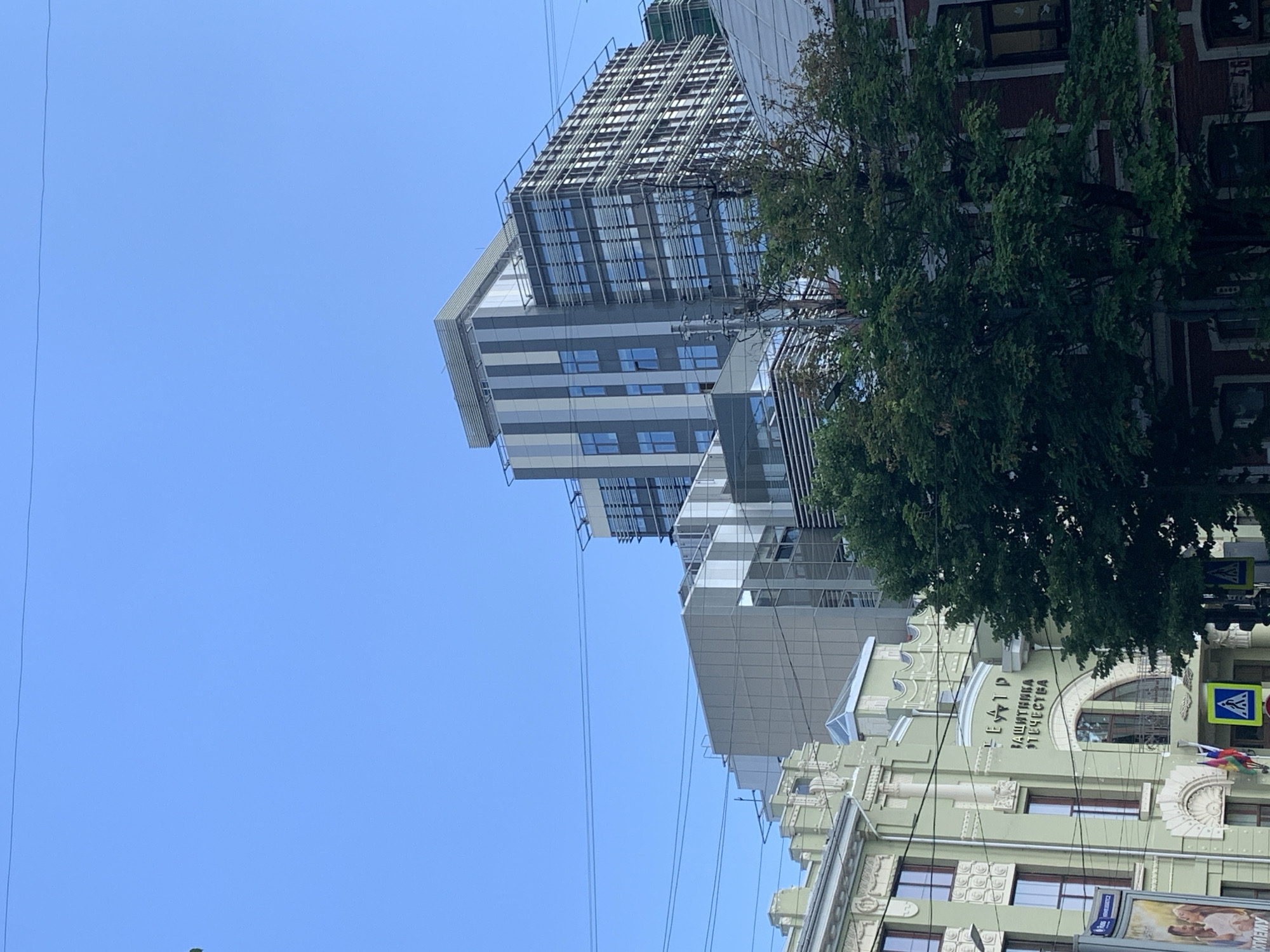 The image captures an urban street scene during the daytime under a clear blue sky. The composition features a mix of architectural styles, dominated by a tall, modern building and an older, more ornate structure.
On the left, a multi-story contemporary building rises, characterized by a facade of light grey and white horizontal panels, interspersed with numerous square windows reflecting the blue sky. The upper sections of this building appear to have an open-frame or lattice-like structure, possibly under construction or a design feature. Several thin power lines or cables are visible against the sky, running horizontally across the frame.
To the right, partially obscured by lush green trees, stands a classical-style building painted in a light green or pale yellow hue, adorned with intricate white architectural details, including decorative pilasters, architraves, and window surrounds. Prominently displayed on the facade of this building is text in Cyrillic: "ТЕАТР ЗАЩИТНИКА ОТЕЧЕСТВА," which translates to "Theater of the Defender of the Fatherland." Below this text, a large arched window is visible. Further to the right, a smaller, darker building with brick-like patterns can be seen.
Additional details include two blue and yellow pedestrian crossing signs (crosswalk signs) visible near the classical building. A billboard with partially visible text, "ЯН...", is located at the bottom right. The overall impression is a bustling urban environment with contrasting architectural elements under bright, clear weather conditions.