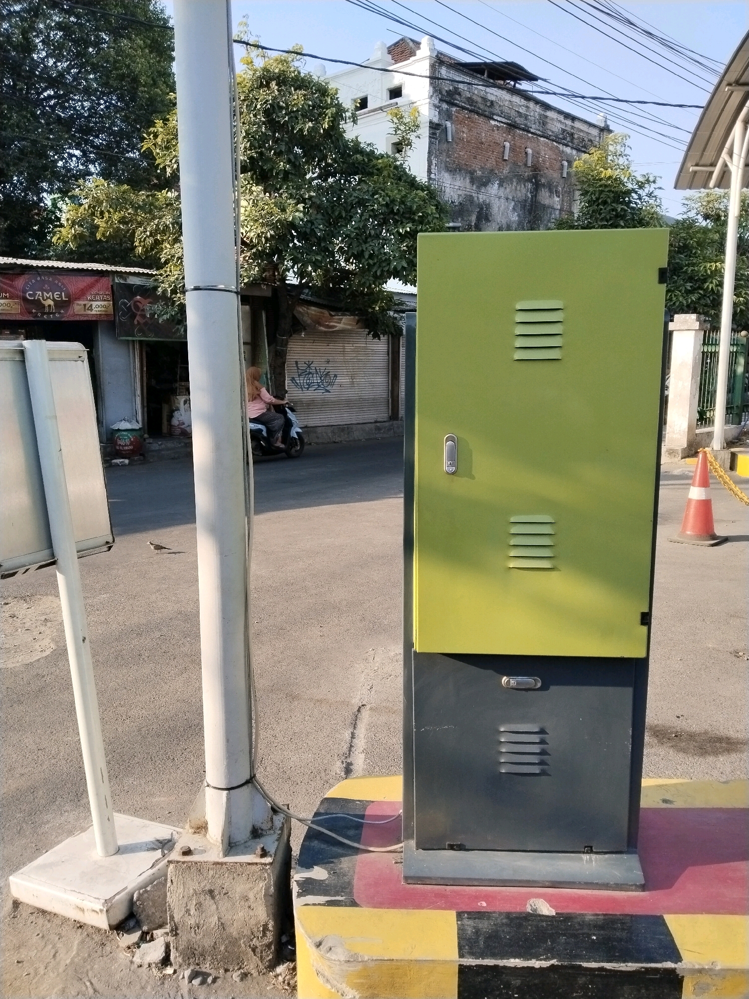 In the city of Wonocolo, Indonesia, a scene unfolds with a prominent lime-green and charcoal-gray utility cabinet positioned on a red, yellow, and black striped border. The cabinet is locked and features horizontal vents on both sections. To the left, a white pole stands, anchored to a concrete base that shows signs of disrepair. Behind the pole, a motorcycle with a rider wearing a light pink shirt and dark pants is visible, heading away from the camera. Adjacent to the street, a building with a distressed brick facade and white trim stands under a bright blue sky, with a tangle of electrical wires overhead. Further left, a storefront with a red awning displaying "CAMEL" text and Indonesian Rupiah prices is partially visible, along with trees and other structures. A small bird is seen on the asphalt in front of the utility cabinet, and a traffic cone with a yellow chain is situated to the right of the cabinet. The ground is asphalt, with some scattered debris.