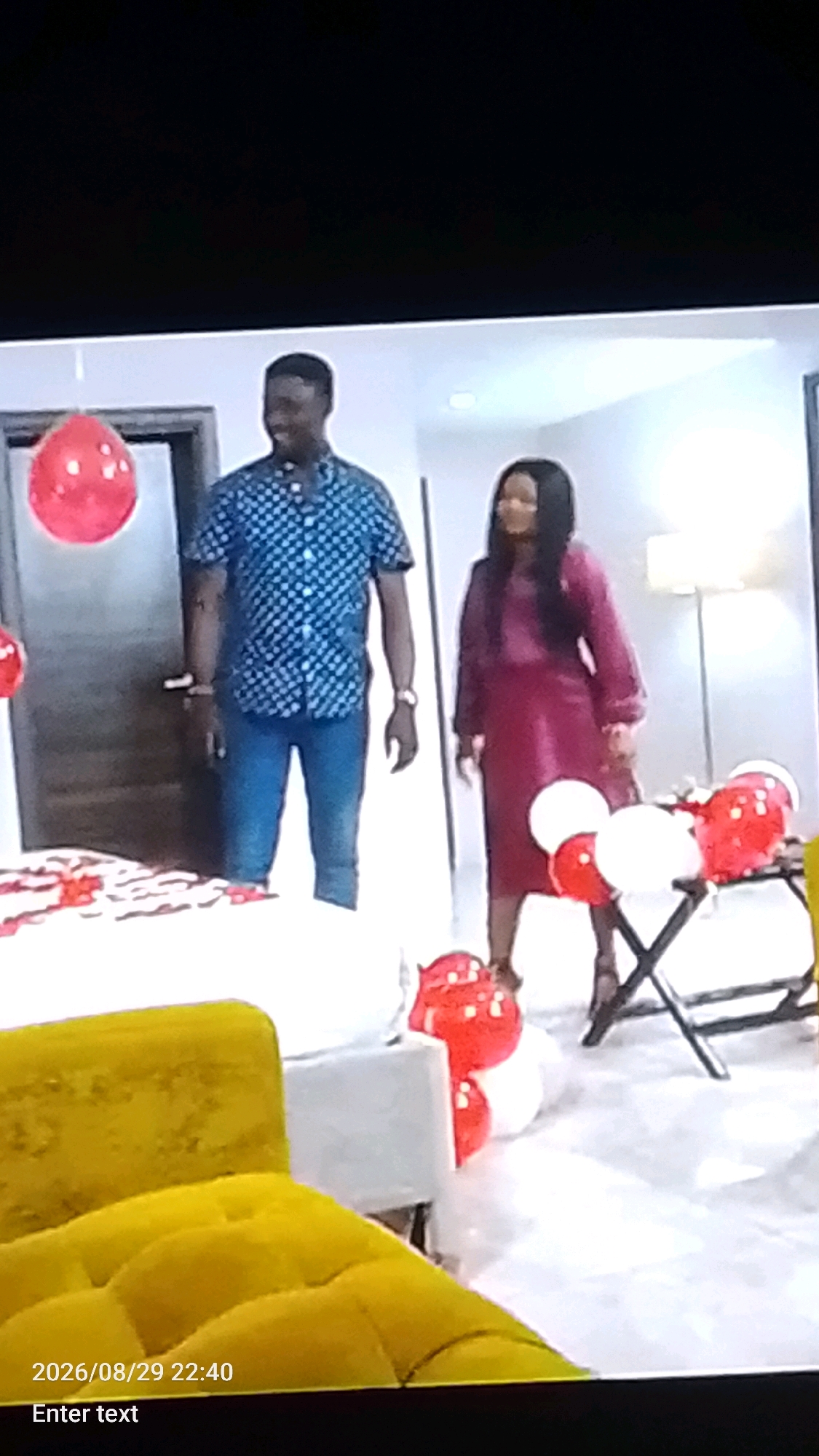 In a domestic setting that appears to be a bedroom in Lokoja, Nigeria, a man and a woman stand near a bed adorned with red rose petals. The man, wearing a blue and white checkered short-sleeved shirt and blue jeans, is positioned on the left. The woman, on the right, is wearing a long-sleeved, deep pink dress. Several red and white balloons are scattered around the room, with some clustered near a folding chair. A portion of a yellow sofa is visible in the foreground. The timestamp "2026/08/29 22:40" is displayed in the bottom left corner of the image.