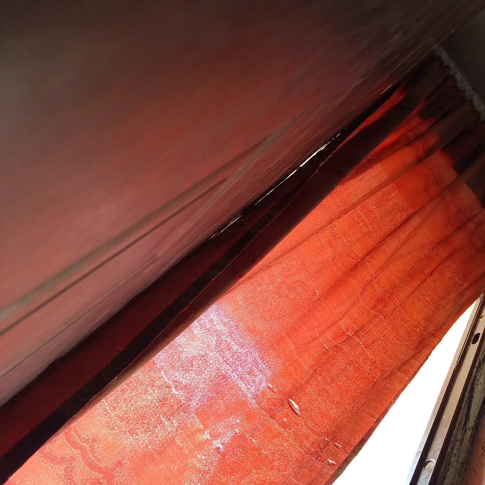 The image shows a close-up of red fabric, possibly curtains or drapery, illuminated by natural light filtering from the side. The fabric has a textured surface and appears to be folded or pleated, creating shadows and highlights. The light source is positioned to the right, casting a warm glow on the fabric. In the background, a dark, indistinct area suggests a confined space, possibly the interior of a room or vehicle. The image is taken at an upward angle, with the fabric filling most of the frame. The setting is a room within a residential building in Jalingo, Nigeria, likely a bedroom or living area.