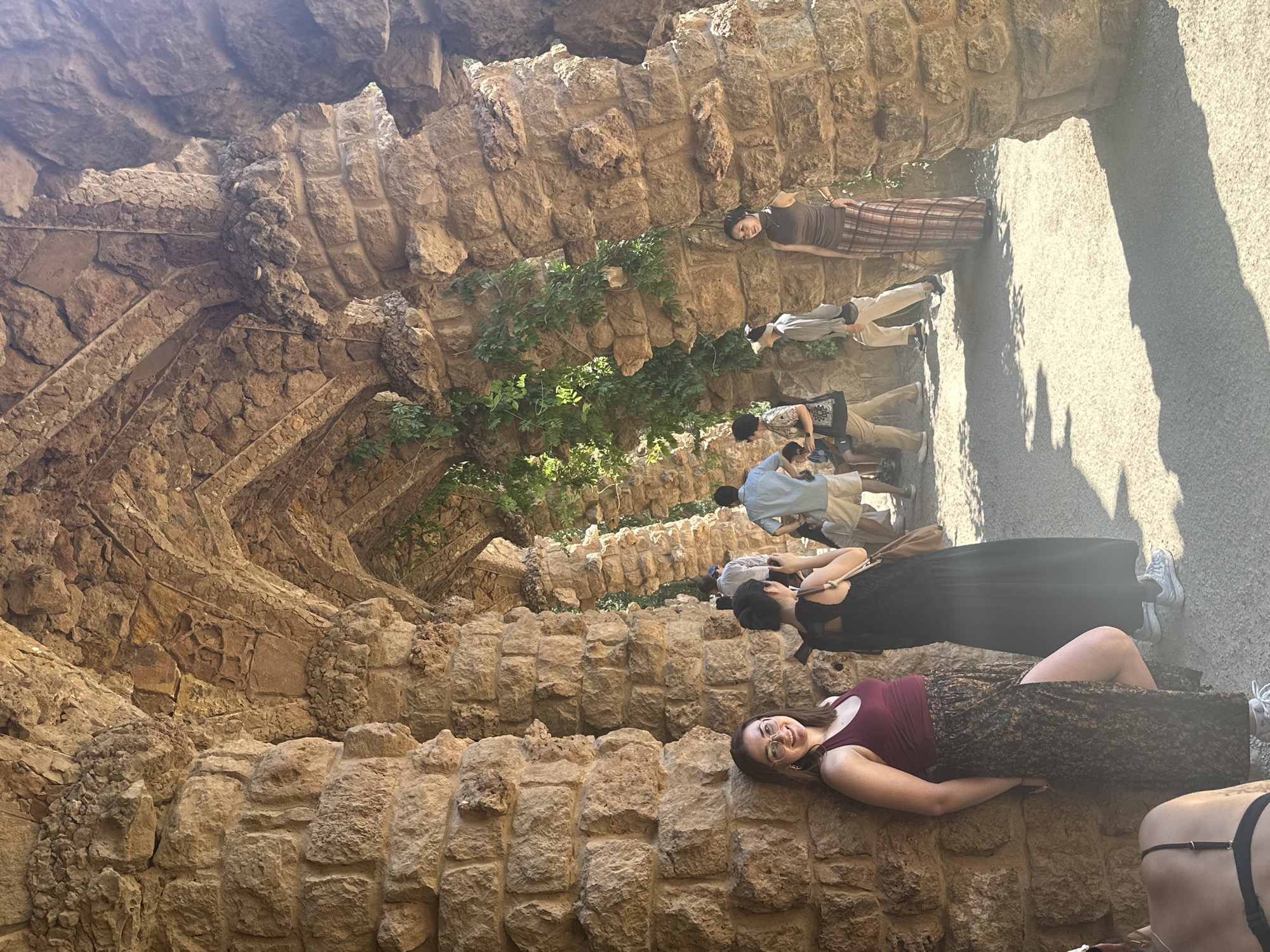This image captures a view of the Portico of the Washerwoman, a well-known architectural element in Park Güell, Barcelona, Spain. The scene is dominated by the distinct, wavy stone columns and arches that form a covered walkway. Sunlight streams through the openings, creating dappled shadows on the sandy ground.
In the foreground, a woman with glasses and shoulder-length brown hair, wearing a maroon tank top and a long, patterned skirt with a high slit, leans against a stone column and smiles at the camera. Behind her, several other individuals are visible. To her right, a woman in a black dress is standing, and further back, a group of people are walking through the portico. These include a man in a light blue shirt and shorts, another person in a light-colored shirt and cap, and a woman in a brown top and a plaid skirt. Greenery vines are draped over some of the stone structures, adding a touch of nature to the architecture.