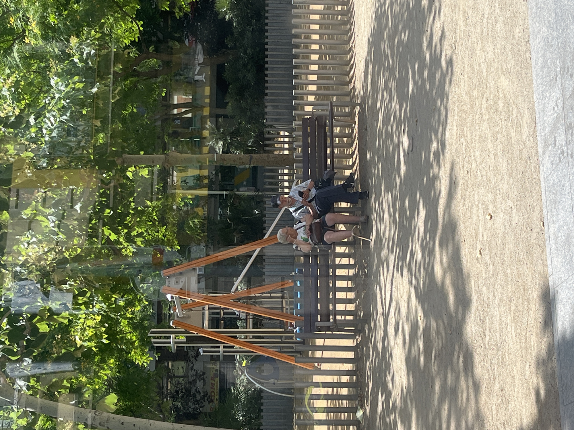 The image depicts a sunny outdoor scene in Barcelona, Spain, likely in a park or public space. In the foreground, the sandy ground is dappled with shadows from overhead trees, suggesting a warm, bright day. A low wooden fence with vertical slats runs horizontally across the middle ground, separating the sandy area from a playground. Behind the fence, a wooden bench is occupied by two elderly individuals. On the left, a woman with short white hair and glasses sits wearing a light-colored top and dark shorts, holding a small bag on her lap and a cane resting beside her. To her right, a man with a cap and glasses is seated, looking down at his phone. He is wearing a plaid shirt and dark trousers. Behind the bench, a children's playground structure with orange A-frame supports and a blue swing is visible. The background is filled with lush green trees and some buildings, indicating an urban environment. The overall impression is one of a peaceful moment in a public recreational area.