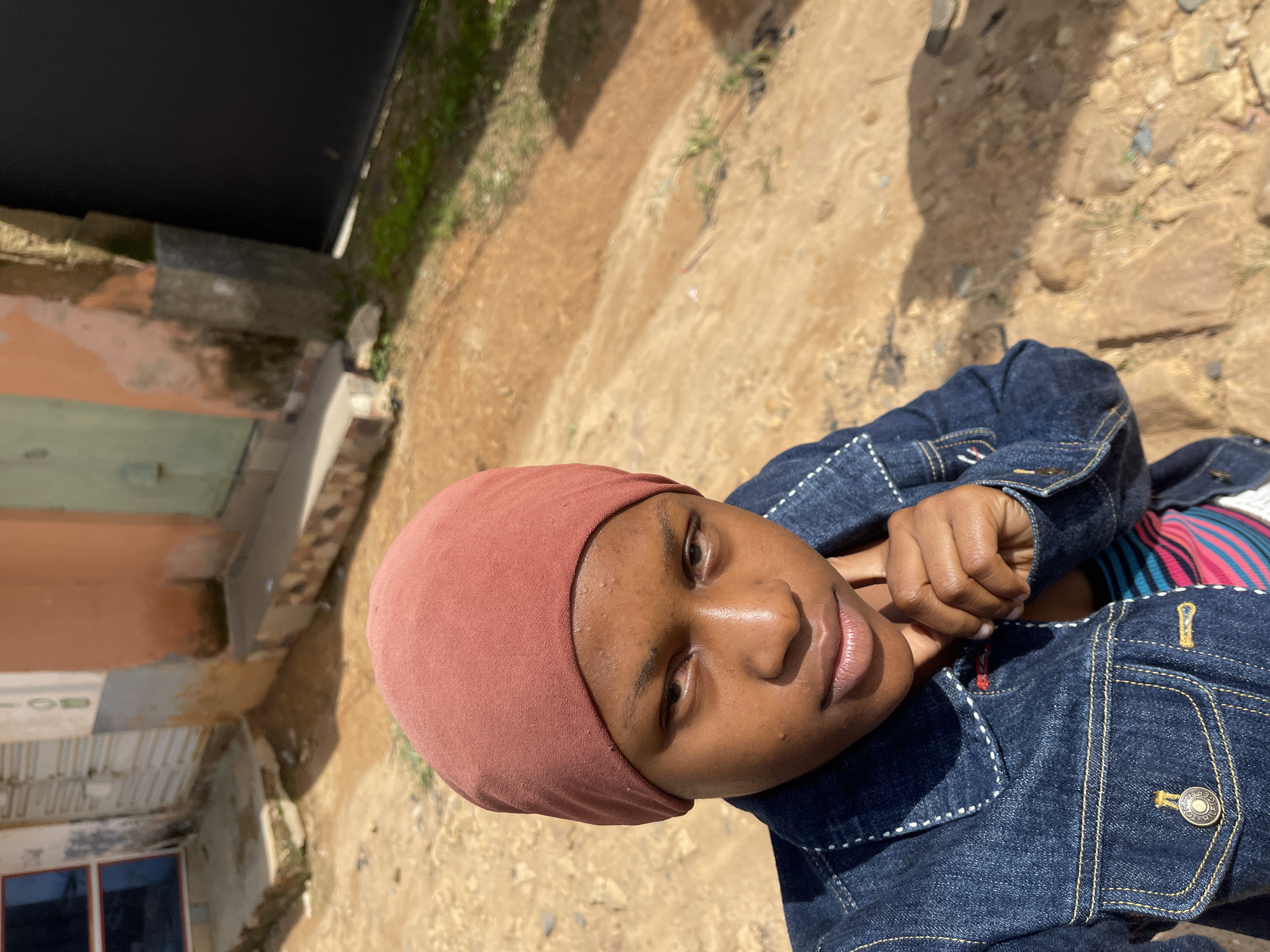 The media file shows a close-up, eye-level, portrait of a young Black person wearing a rust-colored head covering and a denim jacket. The person's face is turned towards the camera, and they are looking directly at the viewer with a neutral expression. Their skin has a warm undertone, and there are some visible blemishes. The denim jacket has white stitching details, and a metallic button is visible on the chest pocket. A bit of striped fabric, with colors of pink, blue, and black, can be seen underneath the jacket.
The background suggests an outdoor setting in a location that appears to be Jos, Nigeria. The ground is a mix of sandy earth and stones, with some patches of dry grass. To the left, there are elements of a building, with peeling paint in shades of orange and green, and corrugated metal roofing. The overall lighting suggests it is daytime, with the sun casting shadows on the ground. The image is captured in a portrait orientation, with the subject's head towards the top of the frame. The scene appears to be a casual, everyday moment, without any specific event being depicted. There is no visible text in the media file.