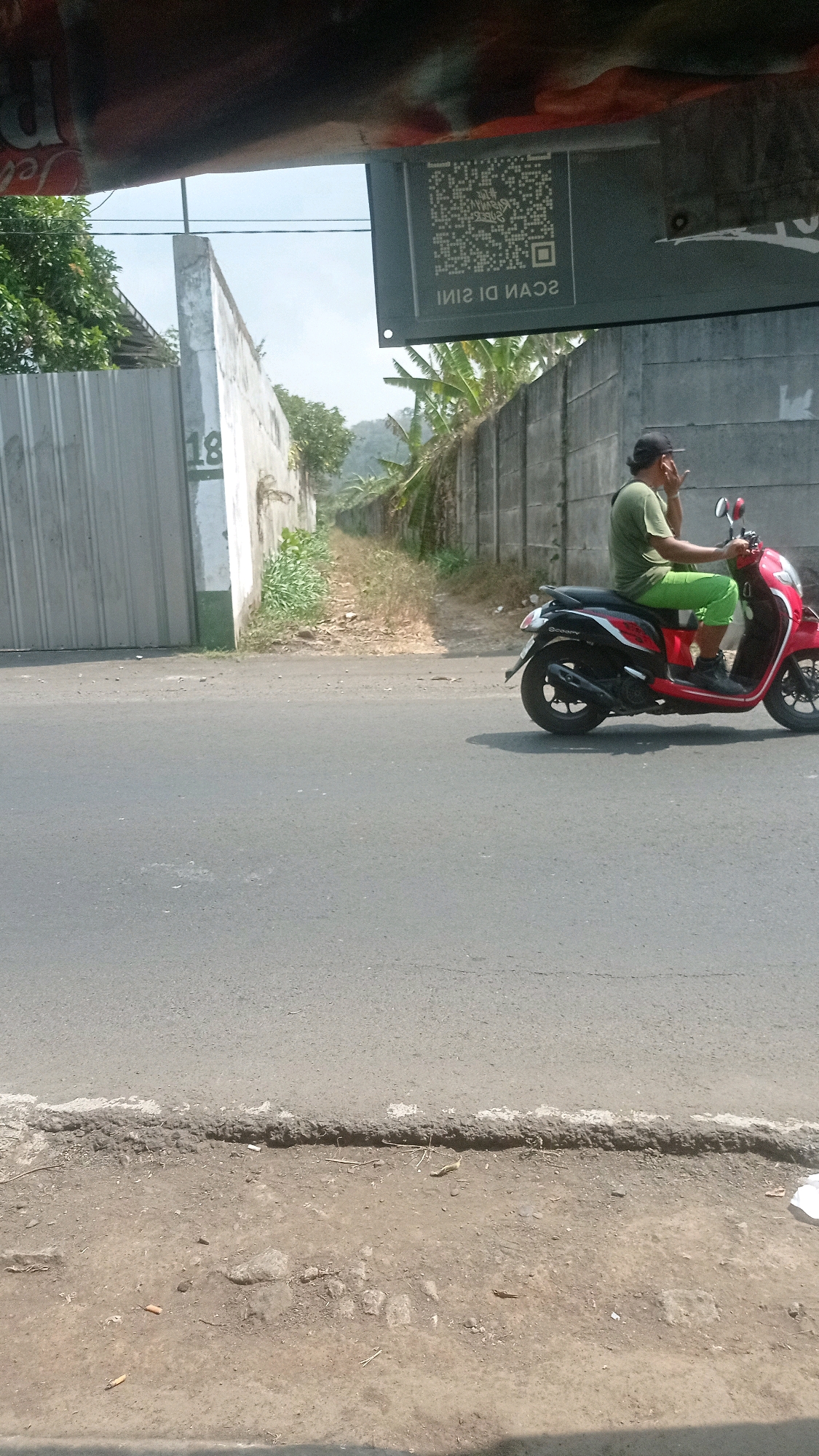 The image shows a street scene in Bedali, Indonesia. A person is riding a red and black scooter down the road, wearing a green shirt and green shorts, and a black cap. The person appears to be smoking a cigarette and is looking to the left. The road is paved and appears to be in moderate condition. To the left of the road, there is a tall, gray metal fence. To the right of the road, there is a concrete wall and some vegetation, including trees and tall grass. Above the wall, a sign with a QR code and text in Indonesian is visible. The sky is overcast. The scene appears to be an ordinary day in a semi-urban or rural area.