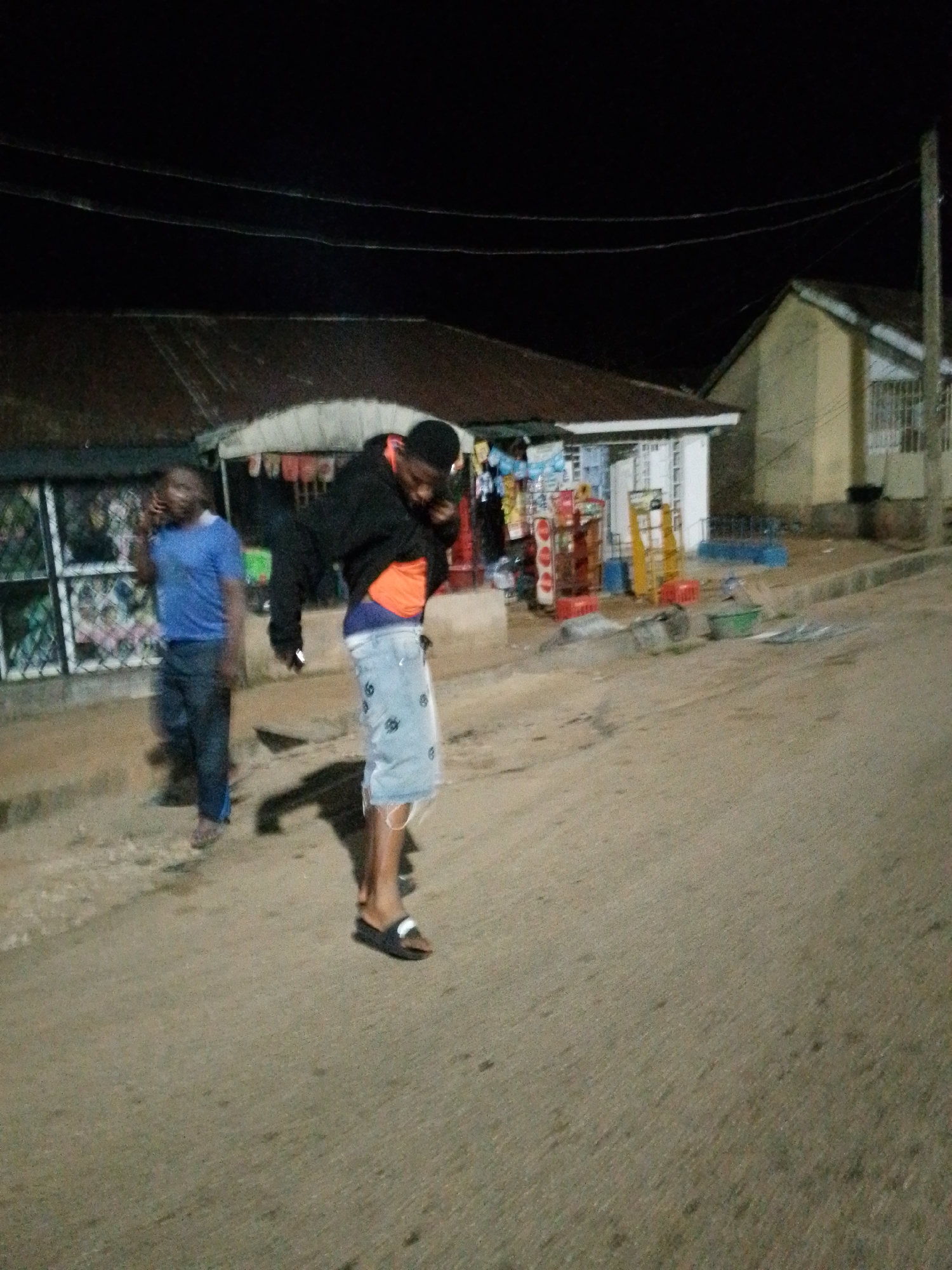 Two men are standing on a dirt road in front of a small shop at night. The man on the left is wearing a blue t-shirt and dark pants and is holding a phone to his ear. The man on the right is wearing a black hoodie, an orange shirt, and denim shorts. He is looking down at his phone, which he is holding in his hand. The shop behind them is lit, and displays various items. The environment is dark, with power lines visible in the background.