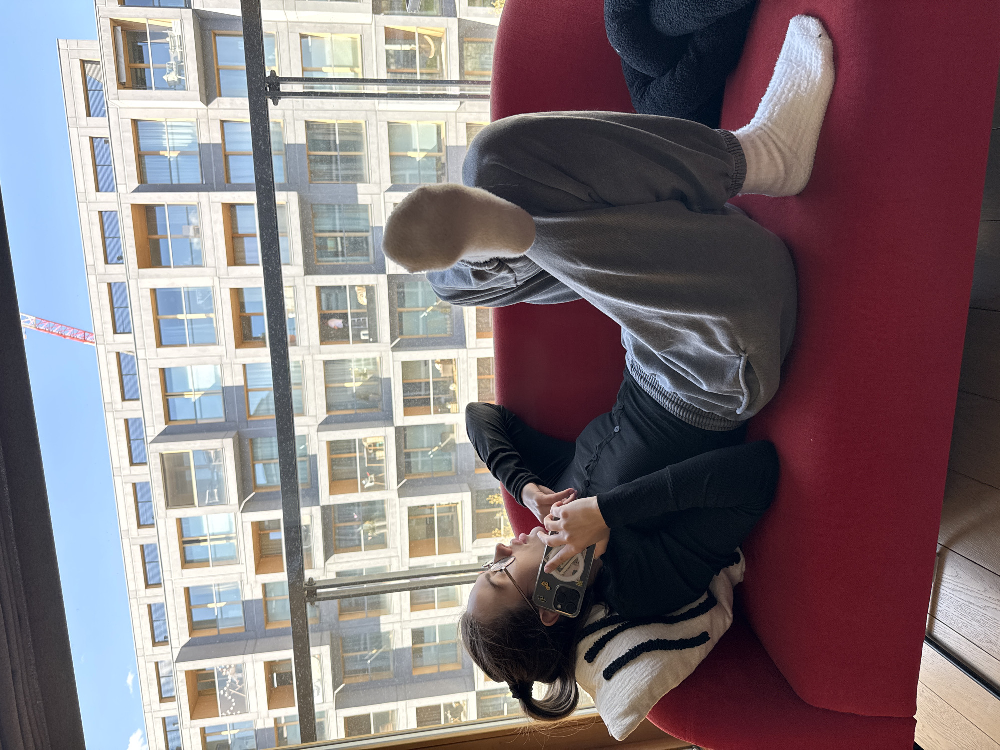 A young woman is shown relaxing on a red sofa, lying on her back with her head resting on a black and white striped pillow. She is wearing a black long-sleeved top, grey sweatpants, and white fuzzy socks. She wears glasses and holds a smartphone with a clear case to her right ear, appearing to be engaged in a phone call.
The scene is set indoors, next to a large window that offers a panoramic view of an urban environment. Outside, under a clear blue sky, stands a modern, light-colored building characterized by numerous square windows. A red construction crane is visible in the distance to the left, indicating ongoing development. The bright daylight suggests it is daytime. The setting is Warsaw, Poland. The phone case has an indistinct design, but no discernible text is legible.
